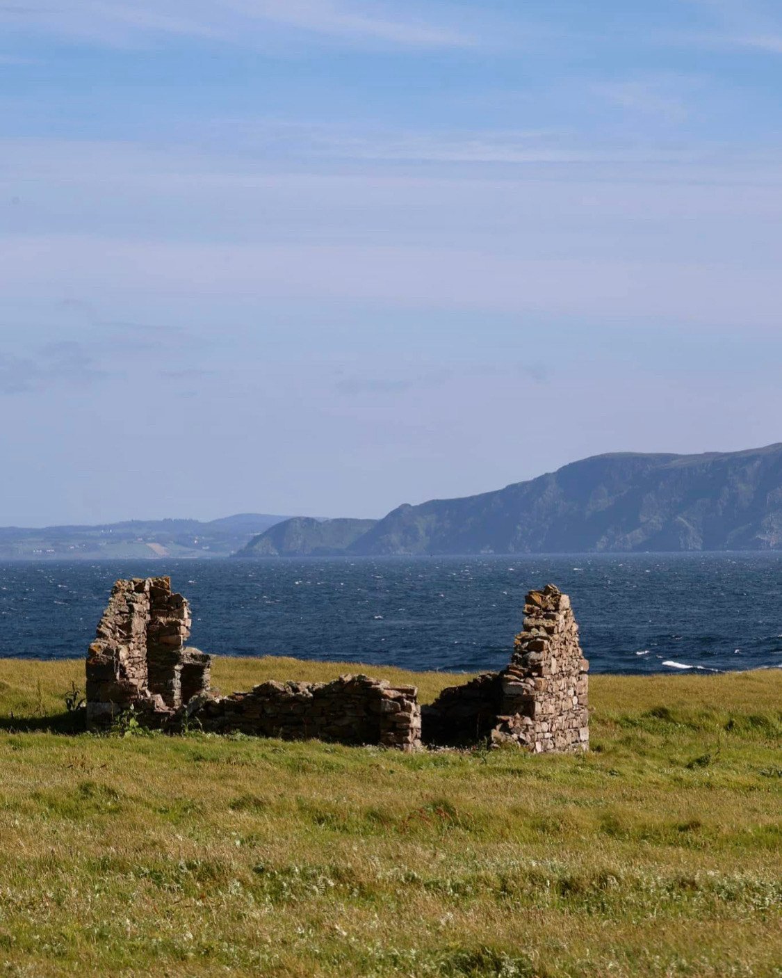 Ruins of an old stone structure on a grassy field near a body of water with mountains in the background under a partly cloudy sky.