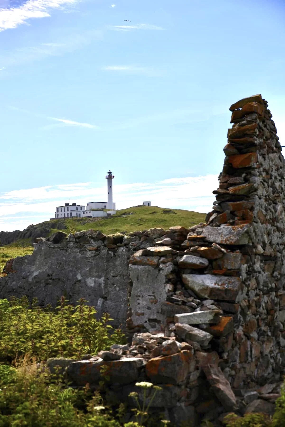 Ruin of stone and concrete structure in foreground, with a lighthouse and a white building on a grassy hill in the background under a blue sky.