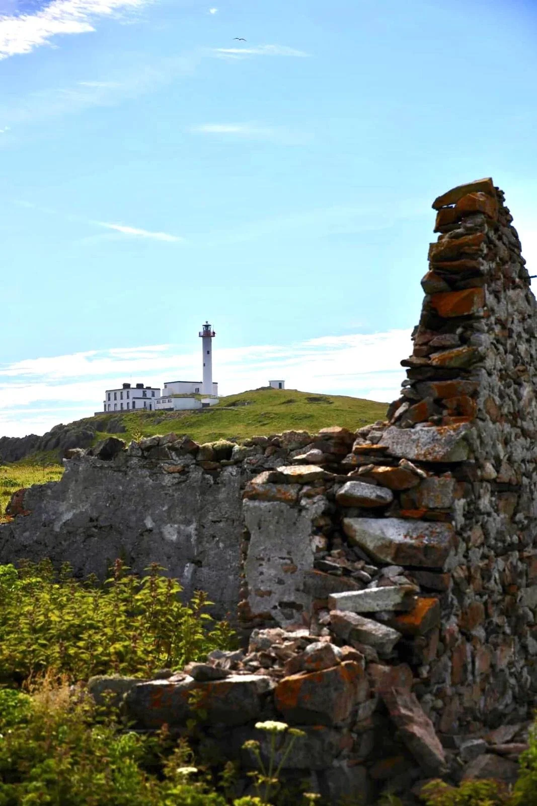 Ruins of an old stone wall in the foreground with a green hill and a lighthouse in the background under a blue sky.