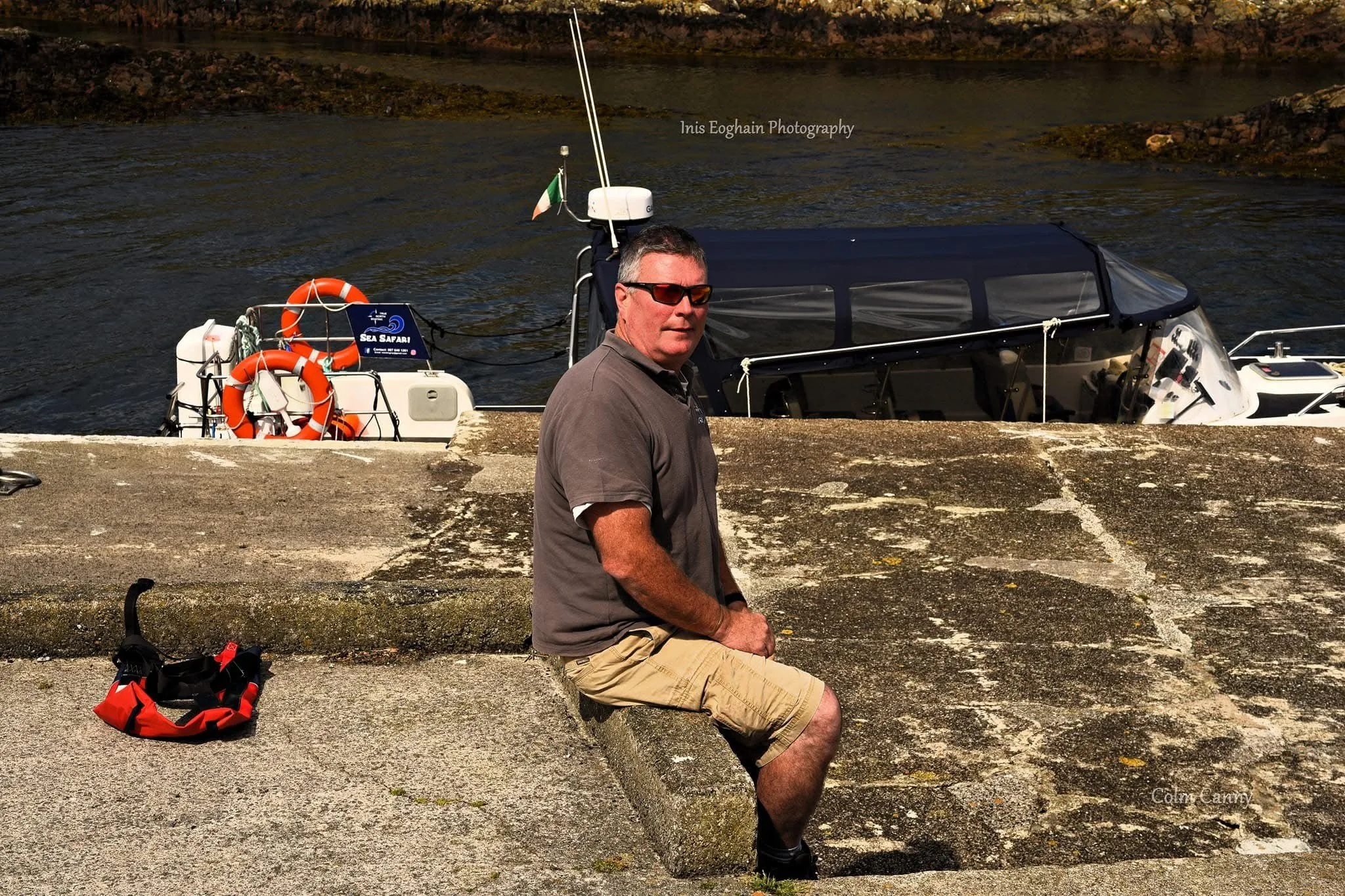 A man sitting on a concrete embankment beside a boat docked in a waterway, with life preservers and equipment on the boat and an Irish flag attached to a mast.