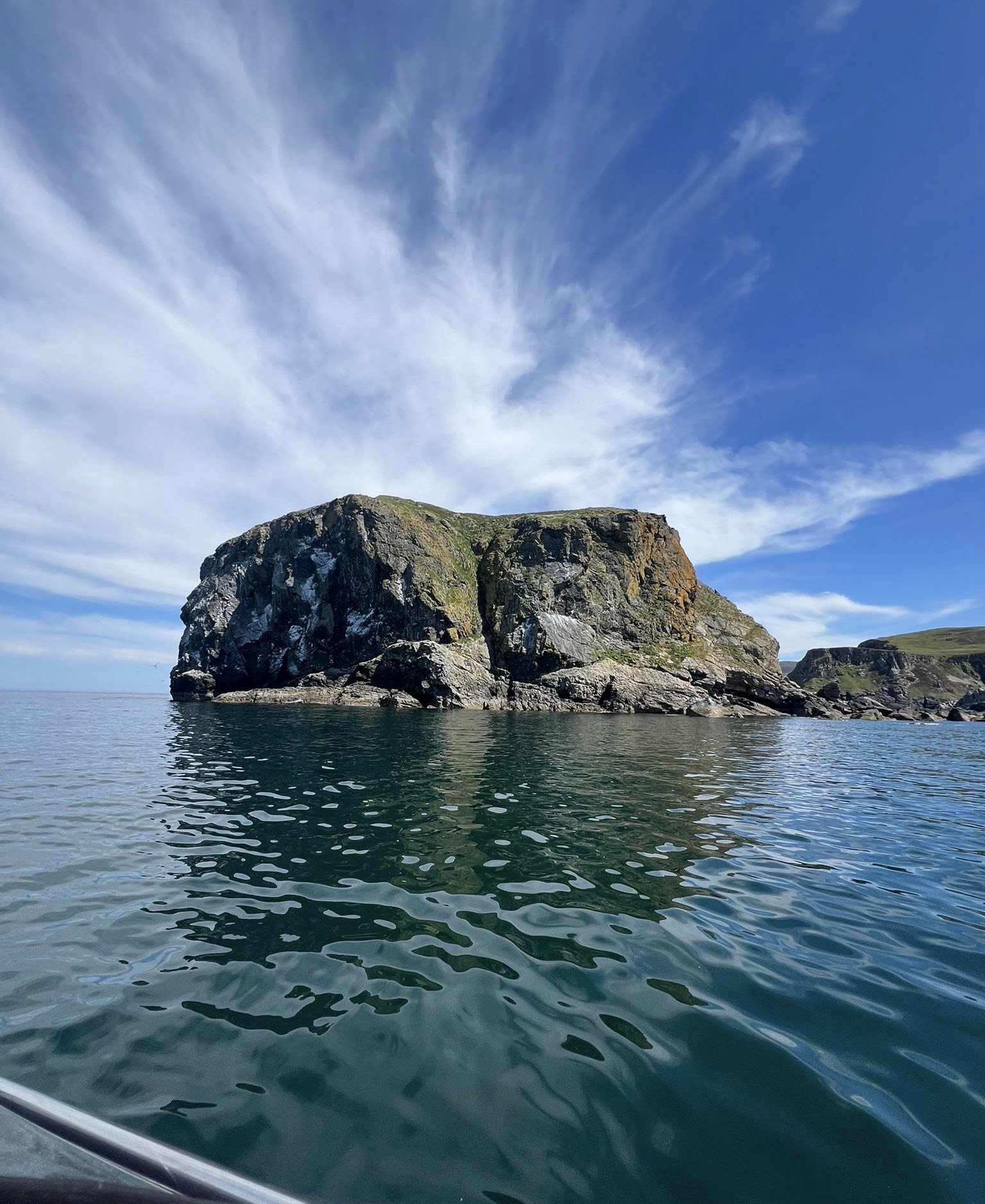 A large rocky island or cliff rising from calm ocean waters under a blue sky with wispy clouds.