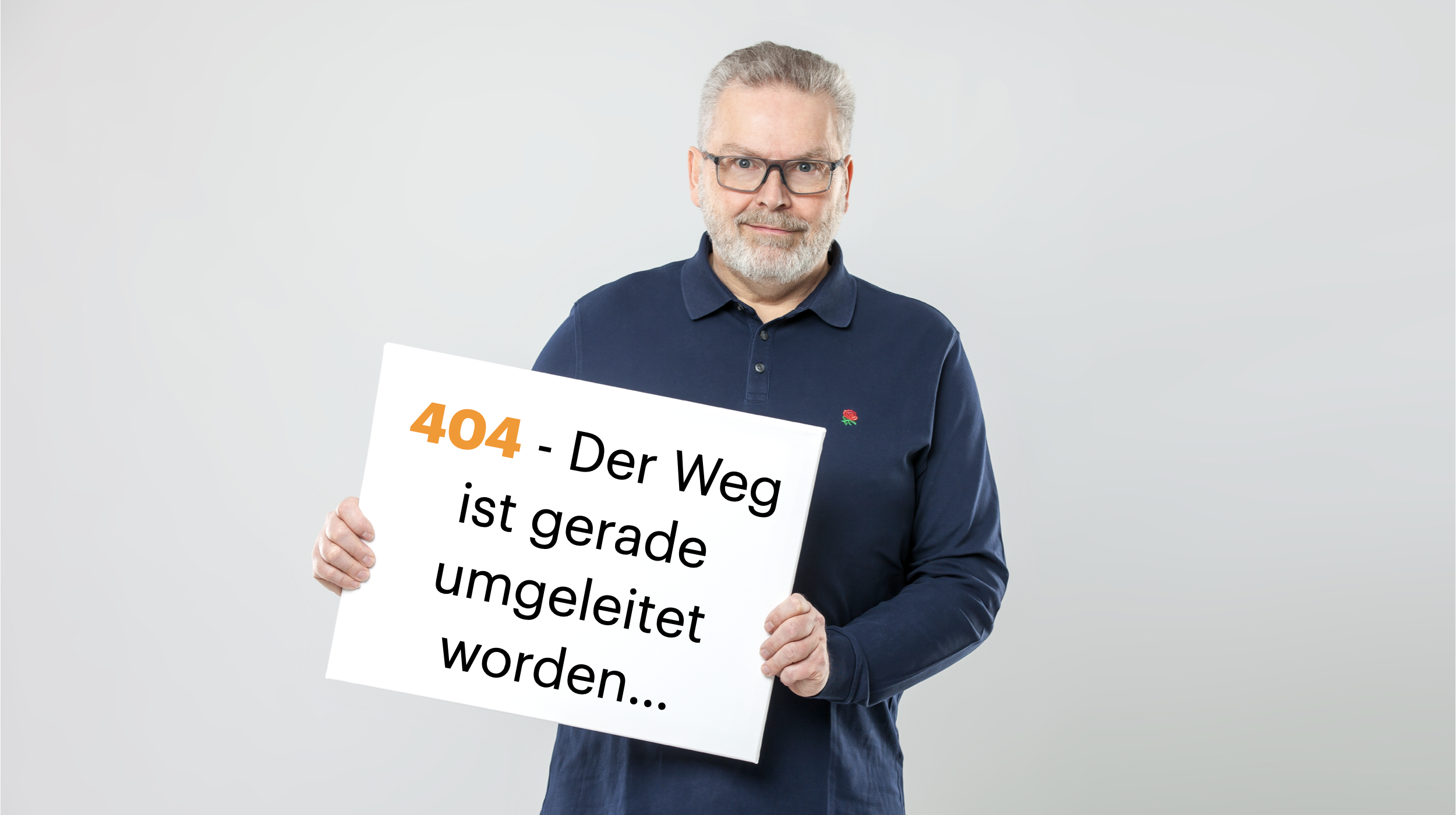 Ein Mann mit Brille und grauem Bart hält ein Schild mit der Aufschrift "404 - Der Weg ist gerade umgeleitet worden...". Er trägt ein dunkelblaues Polo shirt vor hellem Hintergrund.