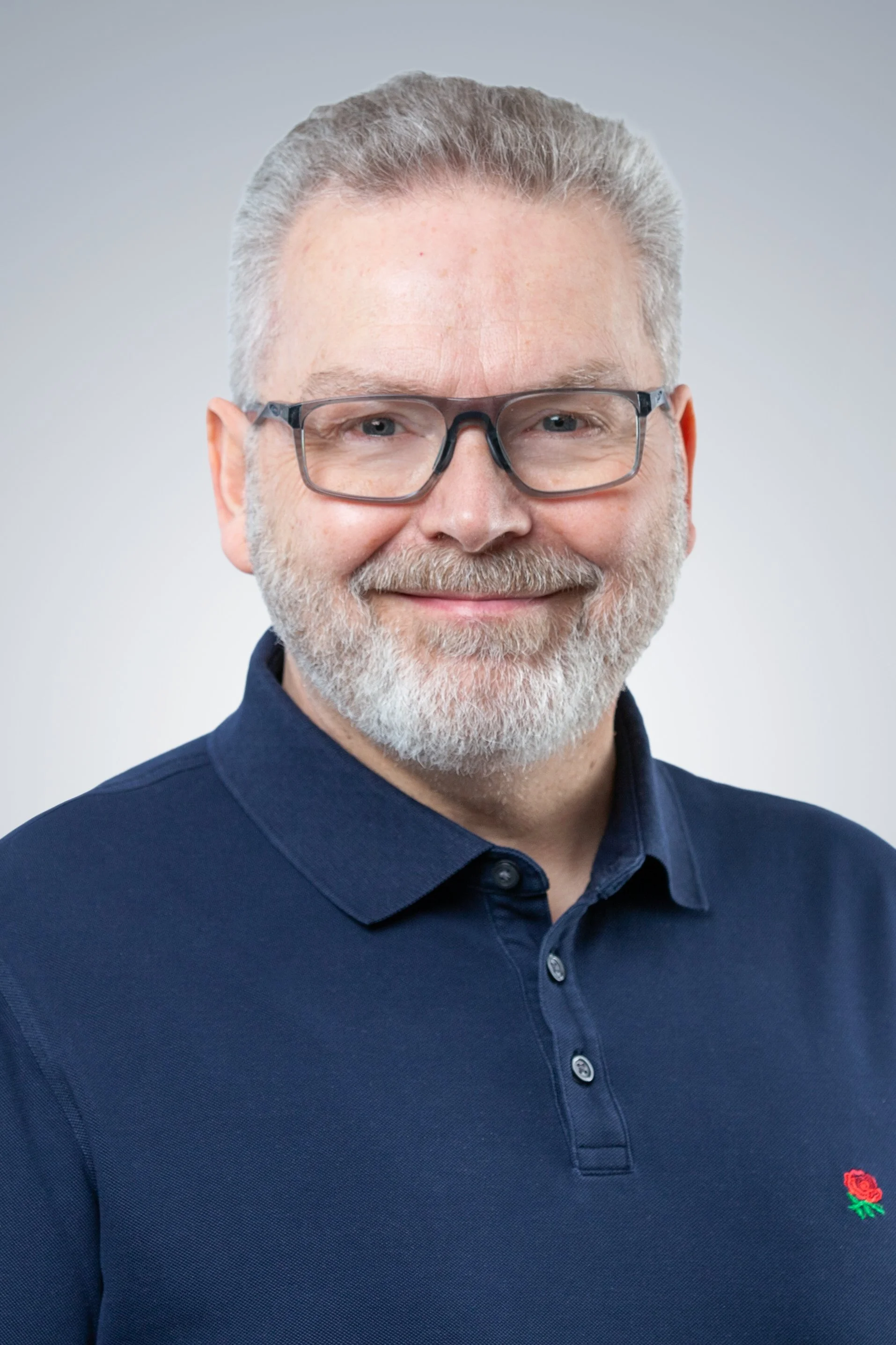Ein älterer Mann mit Brille, grauem Haar und Bart trägt ein dunkelblaues Poloshirt