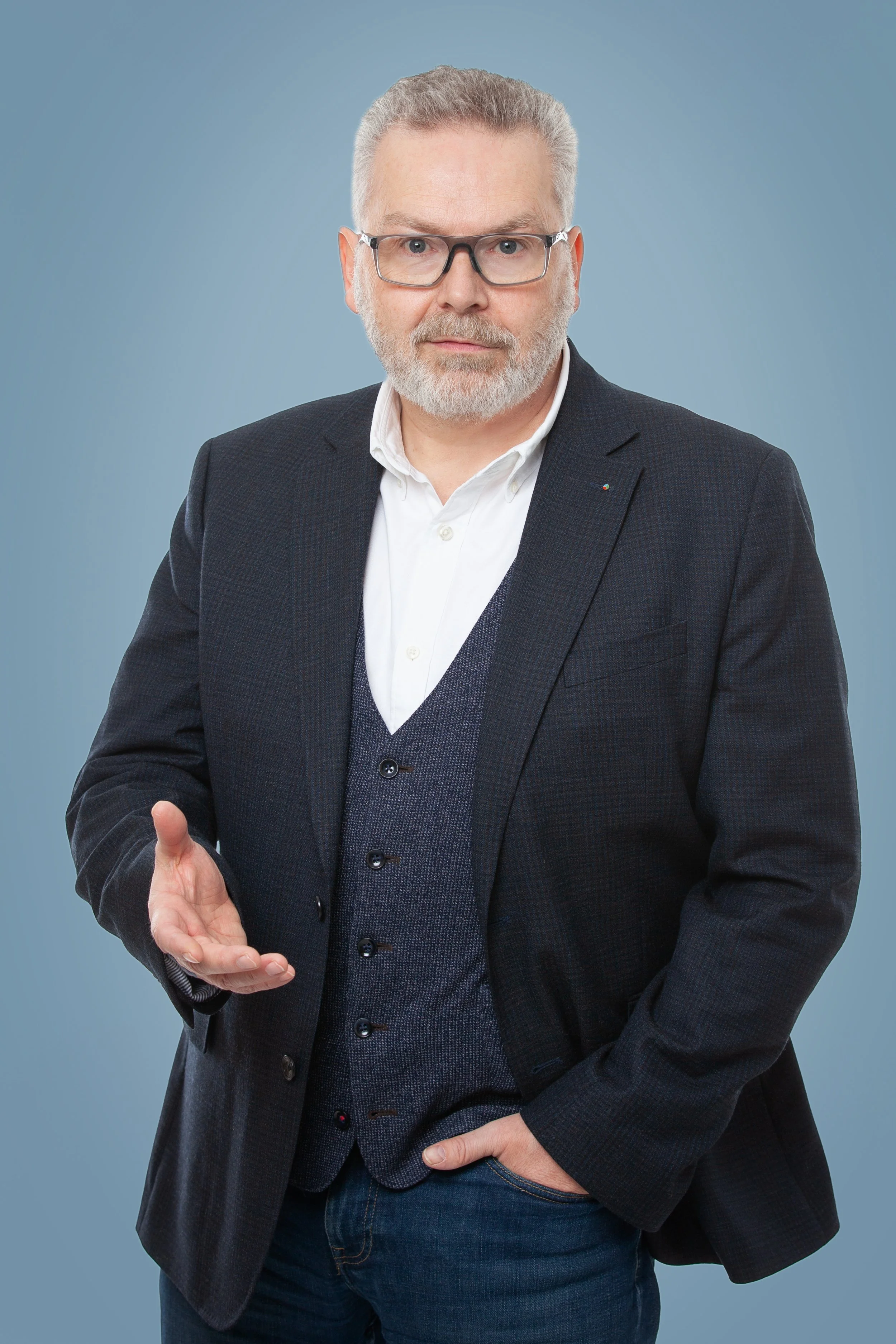 Ein Mann mit Brille, grauem Haar und Bart trägt einen dunklen Anzug und eine weiße Hemd, steht vor einem hellblauen Hintergrund.