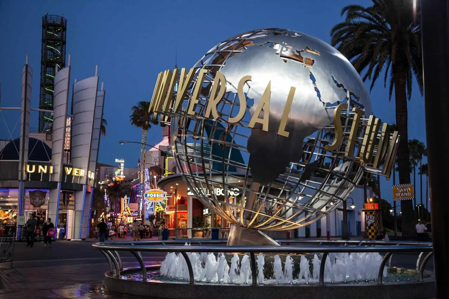 The iconic Universal Studios globe fountain at dusk, with the world map and 'Universal Studios' text, surrounded by palm trees and amusement park buildings illuminated by colorful lights.