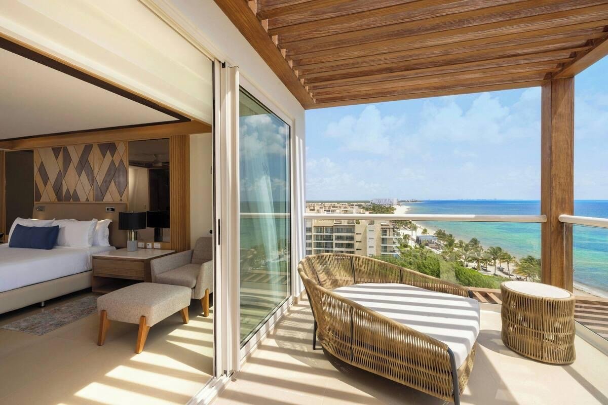 Hotel room view with a sliding glass door open to a balcony overlooking the ocean, featuring outdoor wicker chairs and a clear sky.