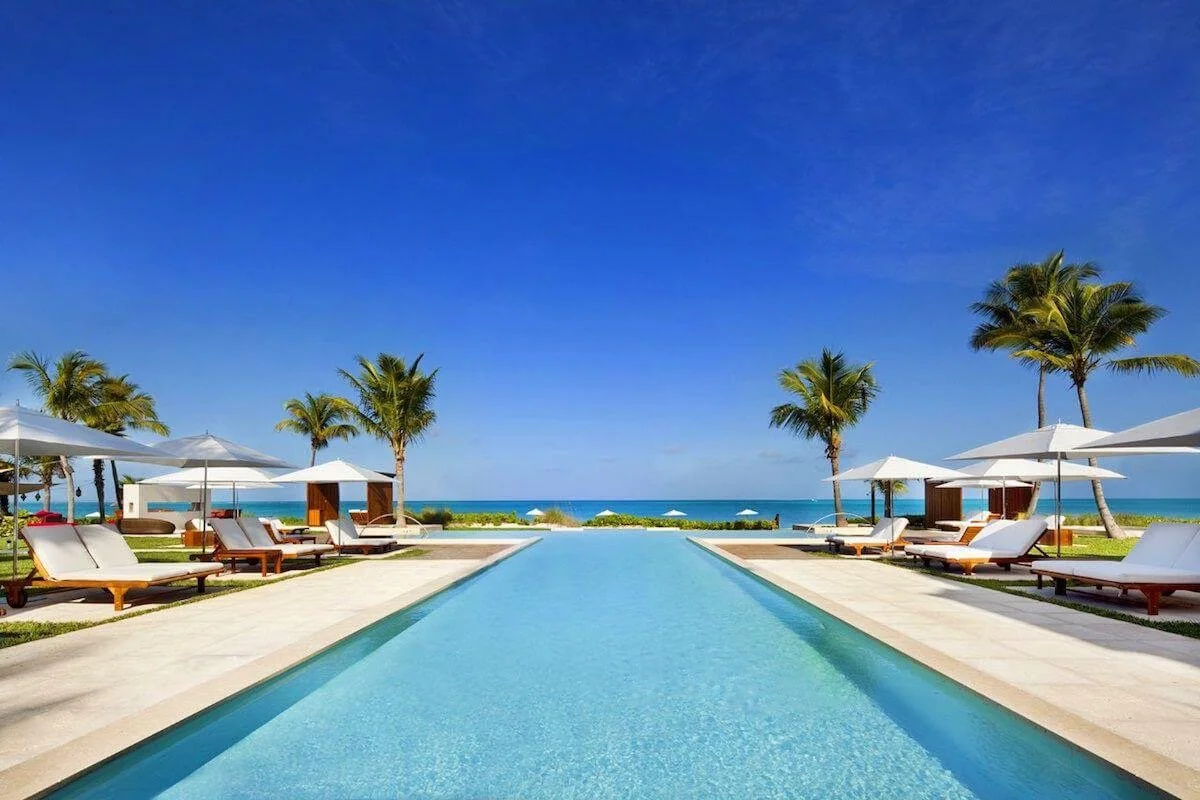Luxury resort swimming pool with white lounge chairs, umbrellas, palm trees, and ocean view.