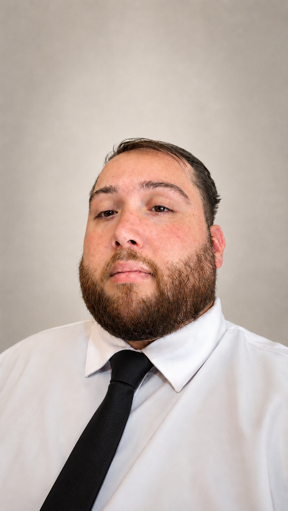 Close-up of a man with a beard wearing a white shirt and black tie, looking seriously at the camera against a plain gray background.