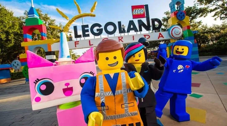 Three people dressed as LEGO characters at Legoland Florida, with Legoland sign in the background, area decorated with colorful LEGO structures.