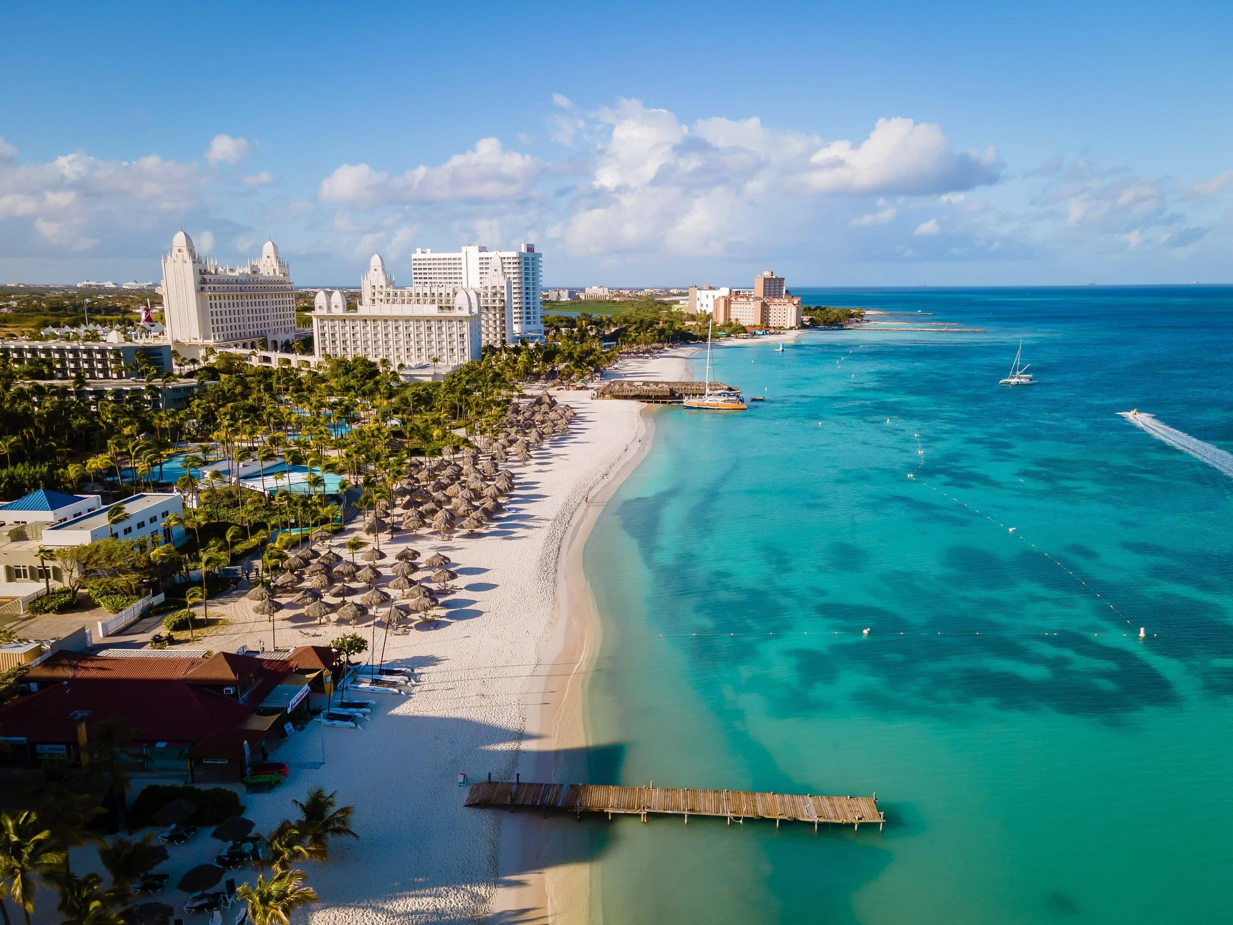 Aerial view of a Caribbean resort with white buildings, palm trees, a sandy beach, and turquoise ocean waters, with boats sailing nearby.
