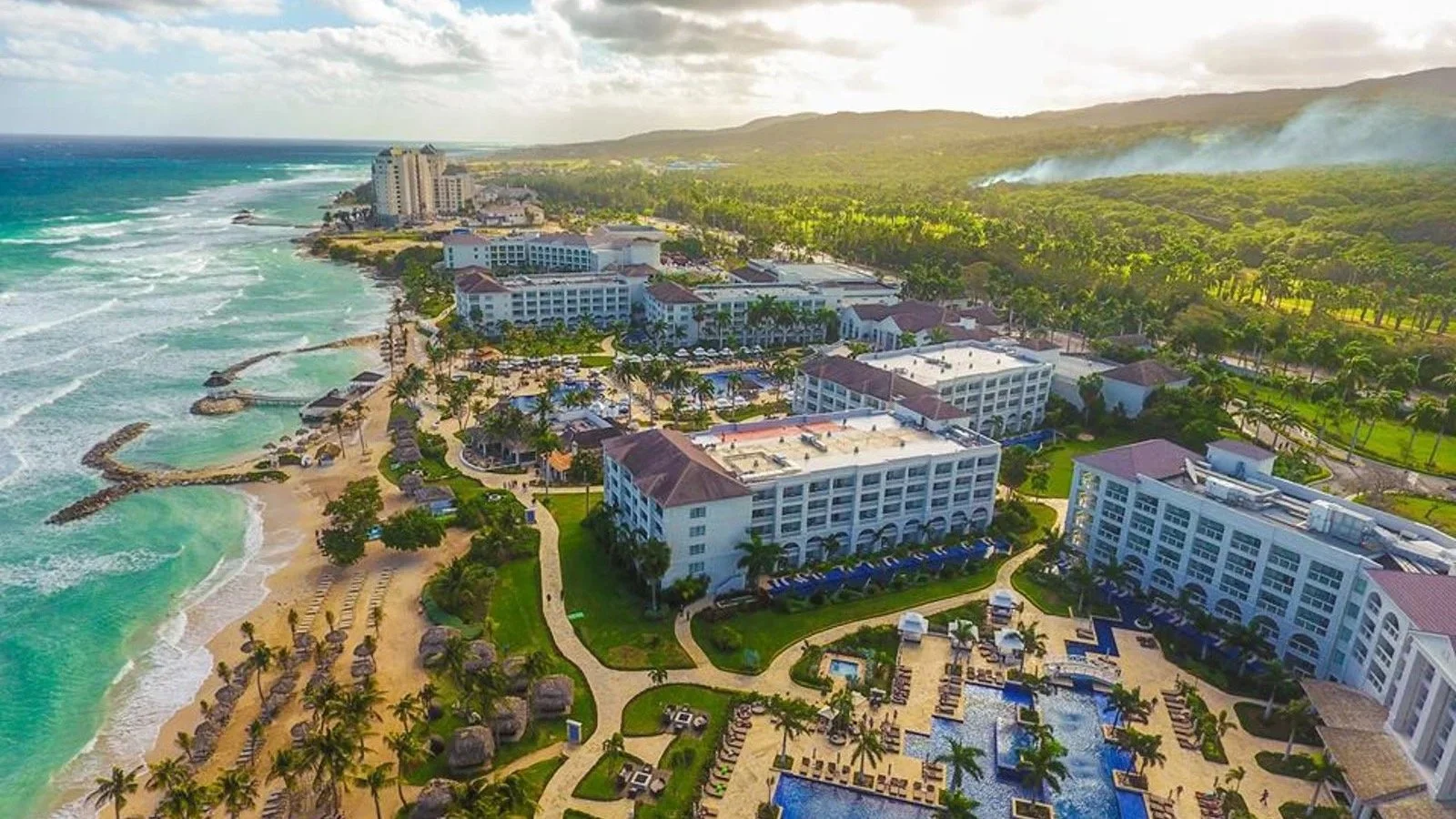 Aerial view of a beach resort with white buildings, pools, and a sandy shoreline lined with palm trees, overlooking the ocean with waves and lush green hills in the background.