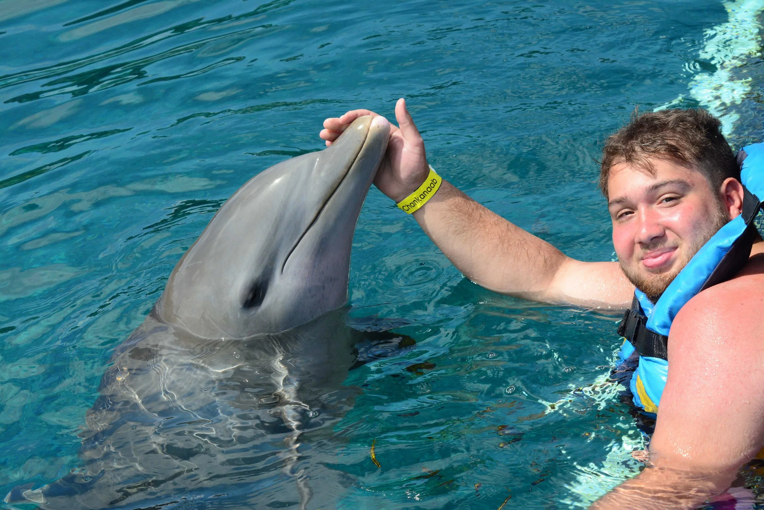 A young man with short brown hair and a beard, smiling, holding a dolphin's bill as it emerges from the water. The background shows blue water, with part of a life jacket visible on the man's shoulder.