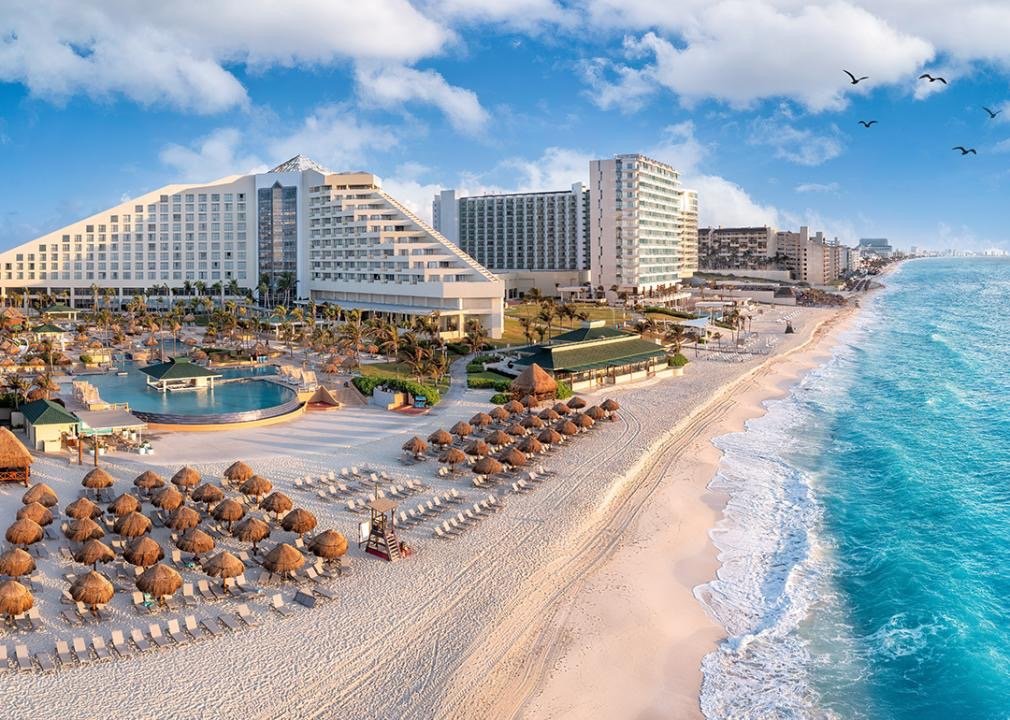 Beachside resort with large hotel building, pool area, straw umbrellas, lounge chairs, and ocean shore with waves and birds in the sky.