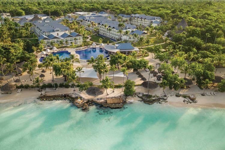 Aerial view of a beach resort with a large swimming pool, palm trees, and turquoise ocean water, surrounded by lush greenery and beachfront properties.