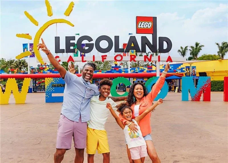 A family of four smiling and celebrating at Legoland California, with large colorful building blocks spelling out 'California' and 'Lego' logos in the background.