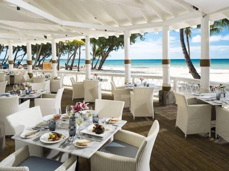 Outdoor seaside restaurant with white tables and chairs, overlooking the beach and ocean, under a white roof with support columns.