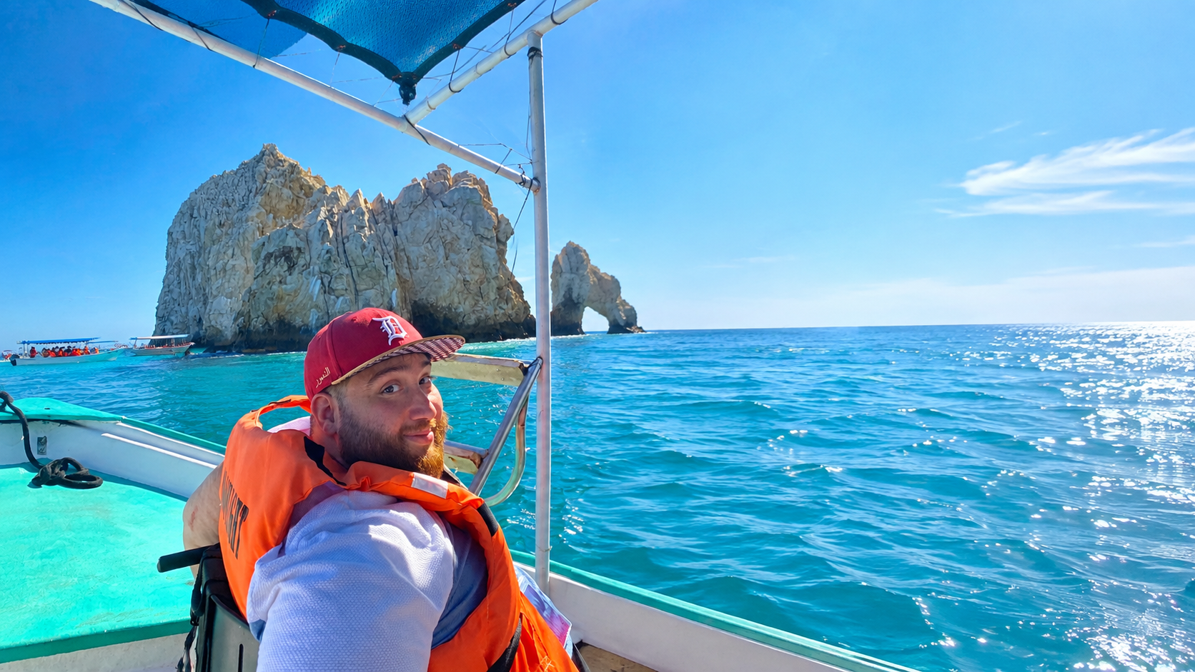 A man on a boat in turquoise waters near large rock formations or small islands, with a clear blue sky in the background.
