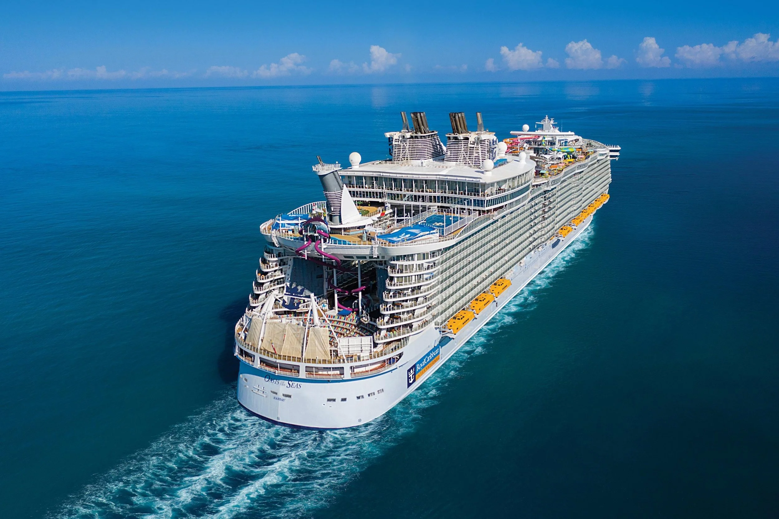 A large cruise ship sailing on clear blue ocean water with a wake behind it, under a bright blue sky with some scattered clouds.