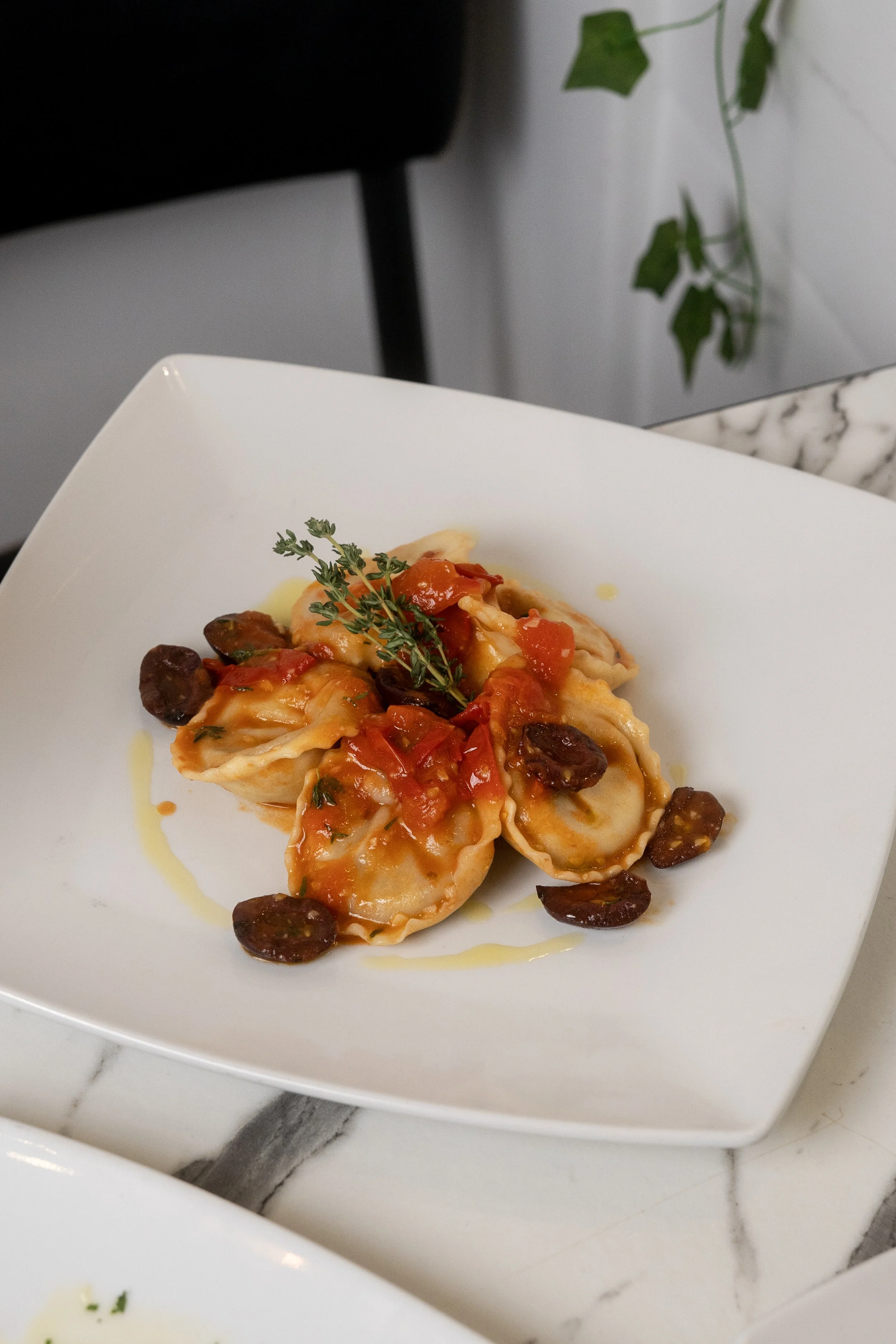Plate of ravioli pasta topped with tomato sauce, olives, and herbs on a white square plate.