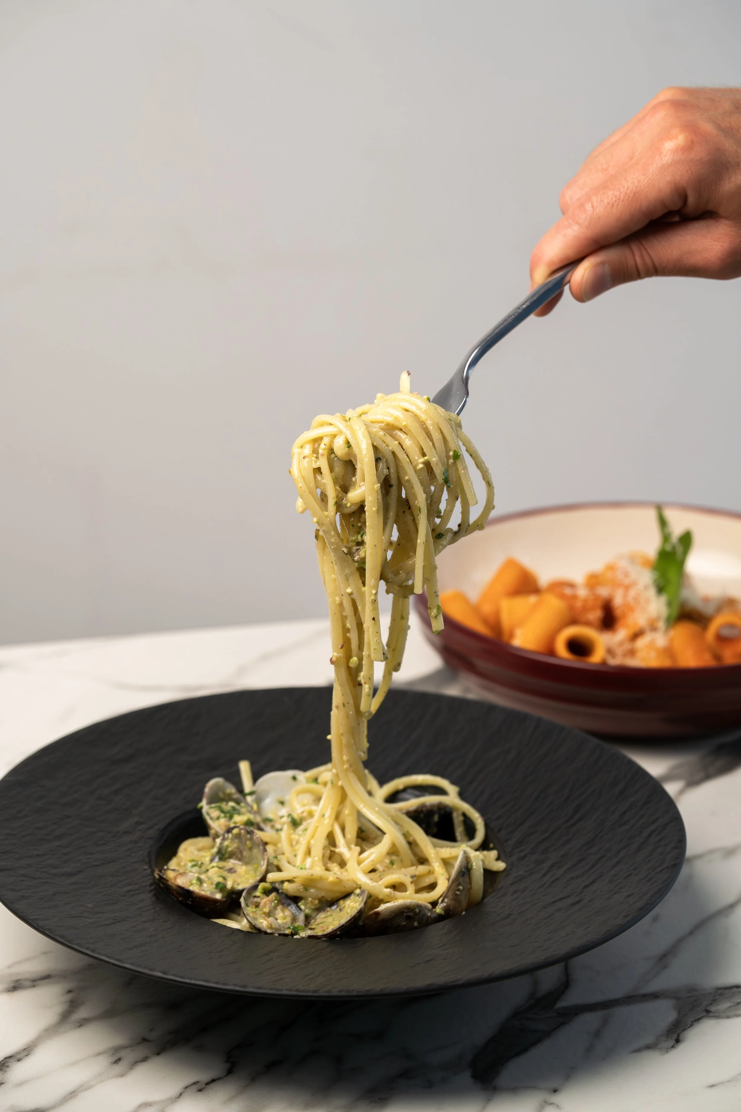 A hand holding a fork lifting spaghetti with clams from a black plate. In the background, there's a red bowl of pasta with orange-colored sauce and garnishes.