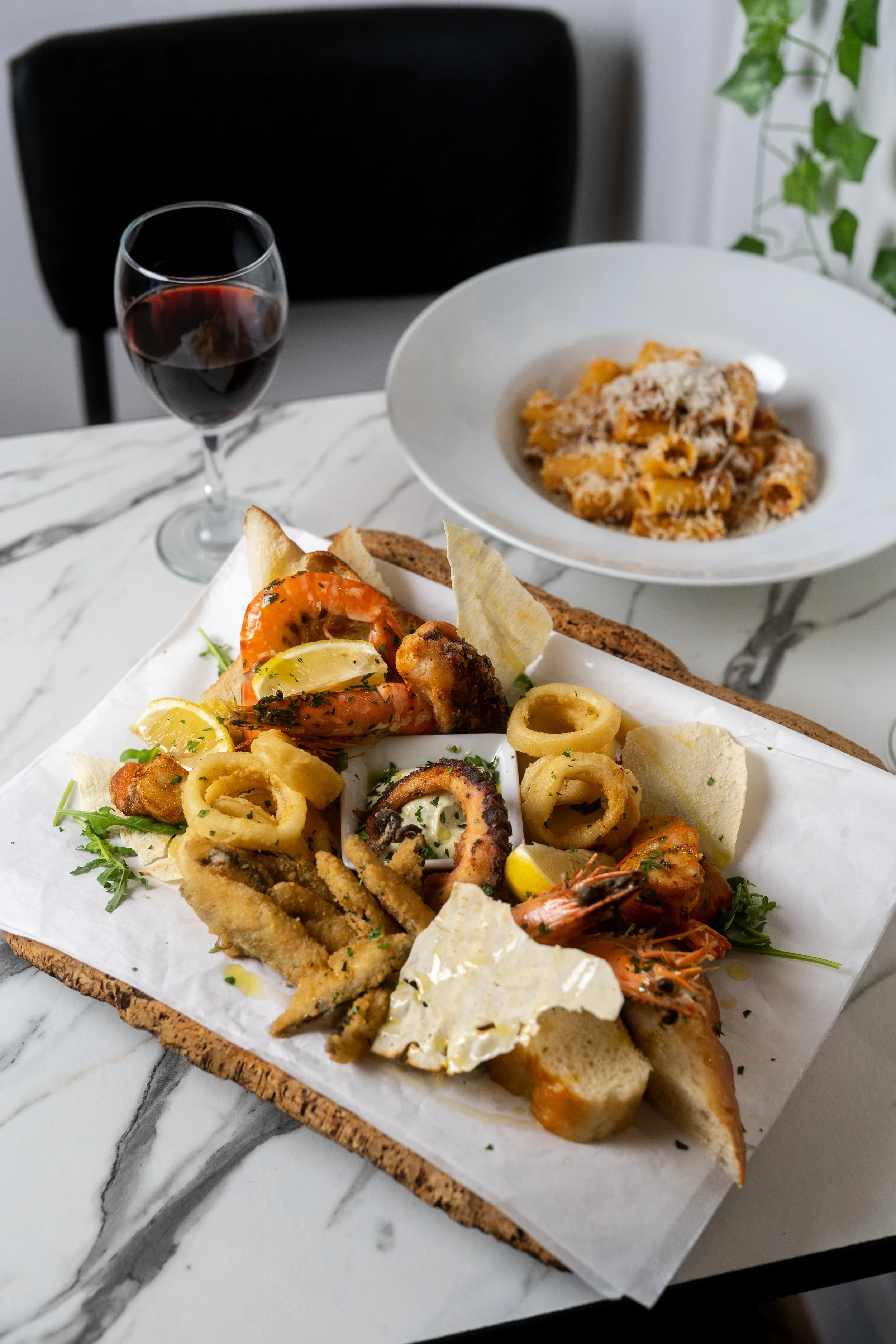 Seafood platter with grilled shrimp, octopus, fried calamari, bread, lemon wedges, and garnishes on a marble table, with a glass of red wine and a plate of pasta in the background.