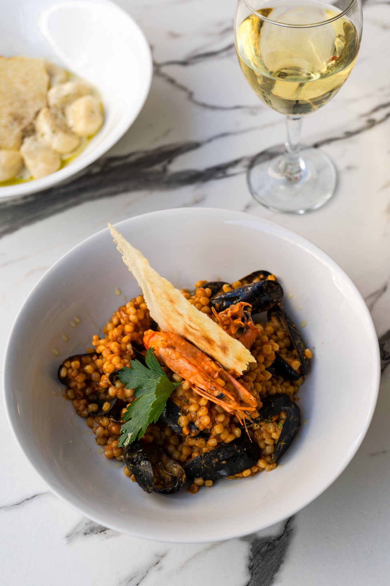 Seafood dish with shrimp, mussels, and lentils garnished with parsley and served with a glass of white wine on a marble table.