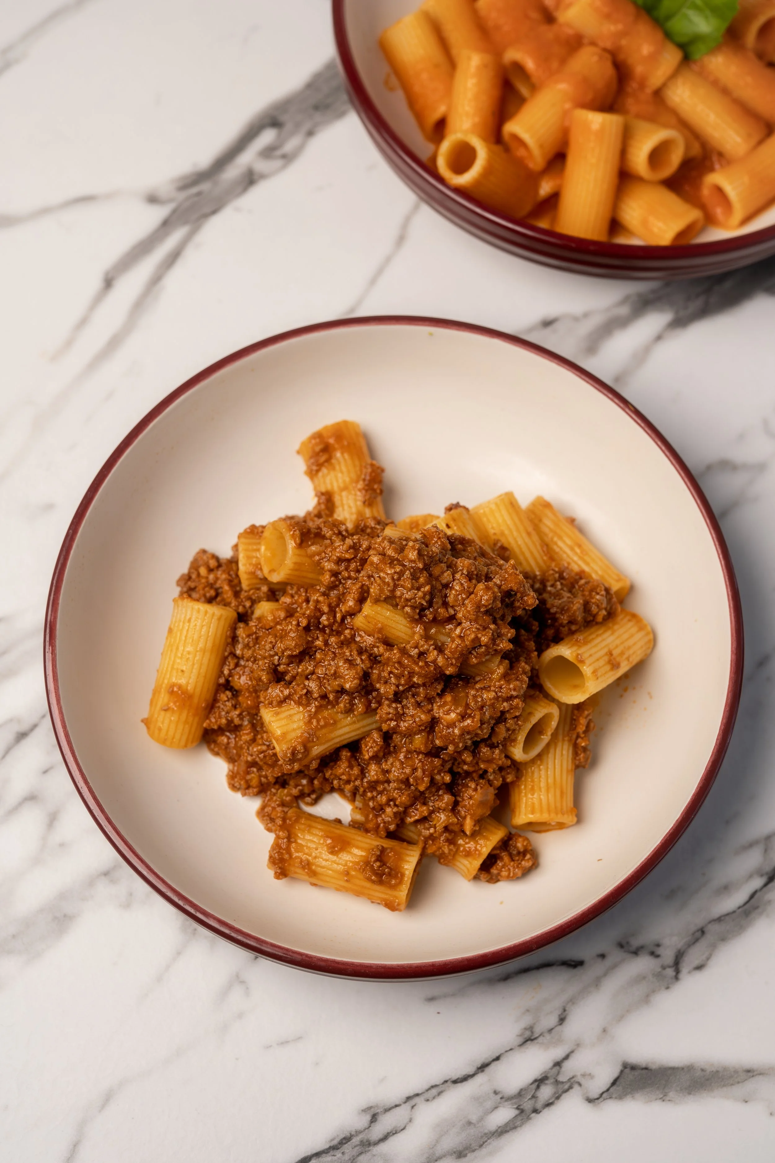 A bowl of cooked rigatoni pasta topped with Bolognese meat sauce on a white marble surface, with another bowl of pasta in the background.