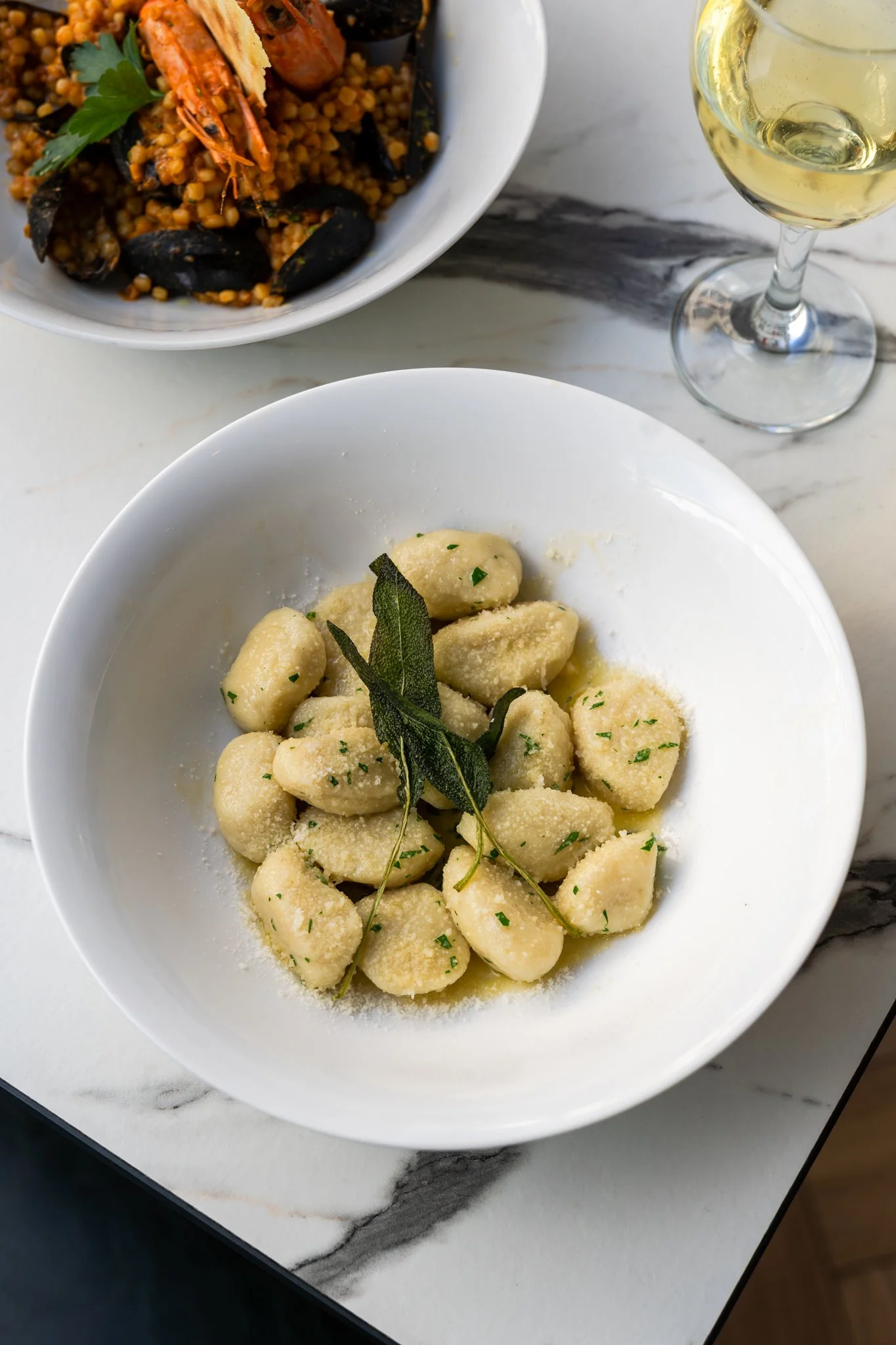 A bowl of gnocchi garnished with sage leaves and chopped herbs, with a bowl of seafood paella and a glass of white wine in the background.