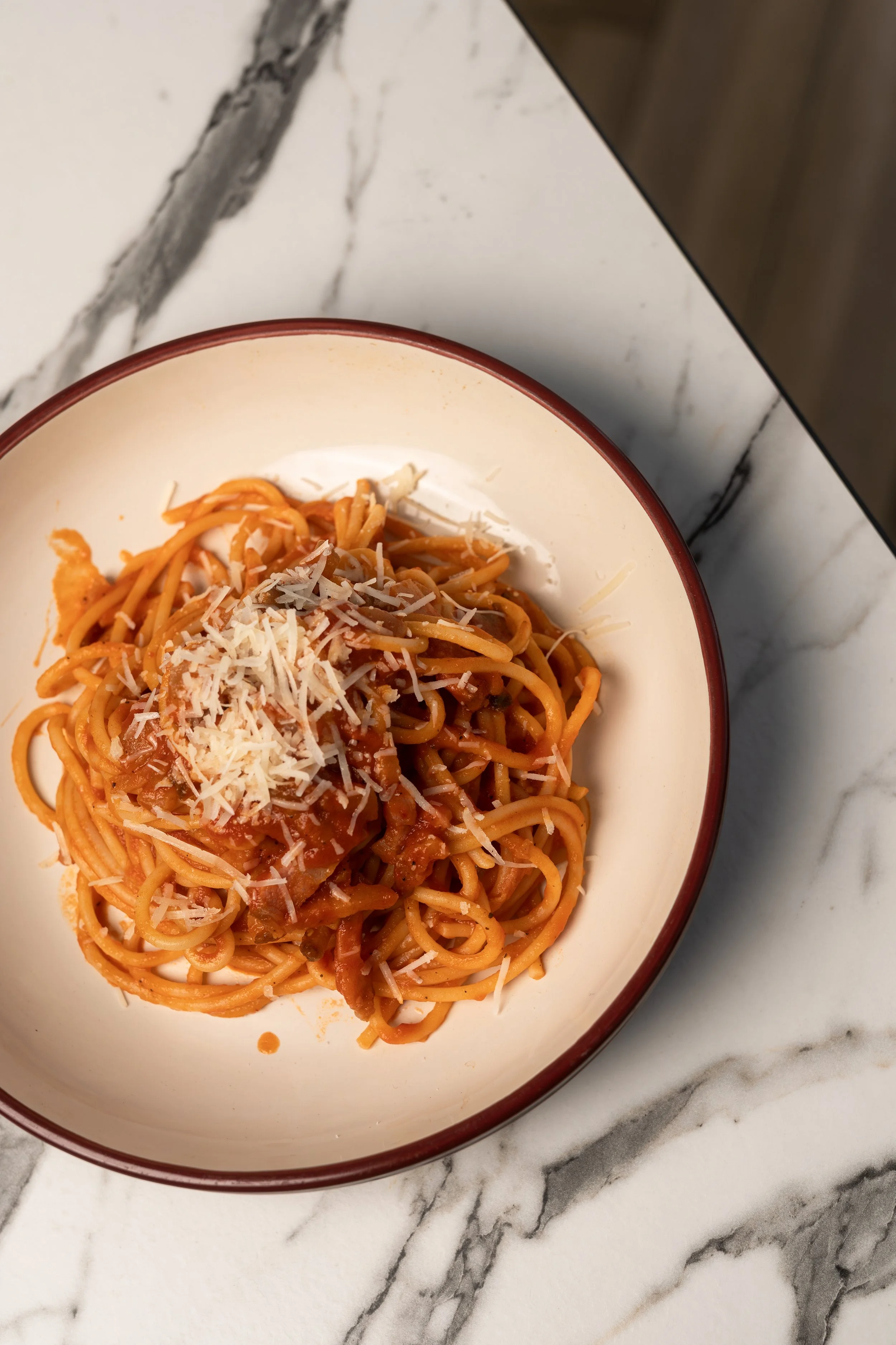 A bowl of spaghetti pasta with tomato sauce and grated cheese on top, placed on a marble countertop.