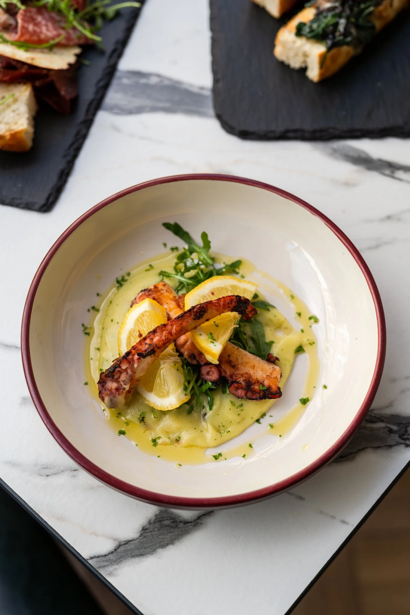A bowl of grilled octopus tentacle garnished with lemon slices, placed over a bed of green salad and yellow sauce, on a white marble surface with slate serving platters of sandwiches in the background.