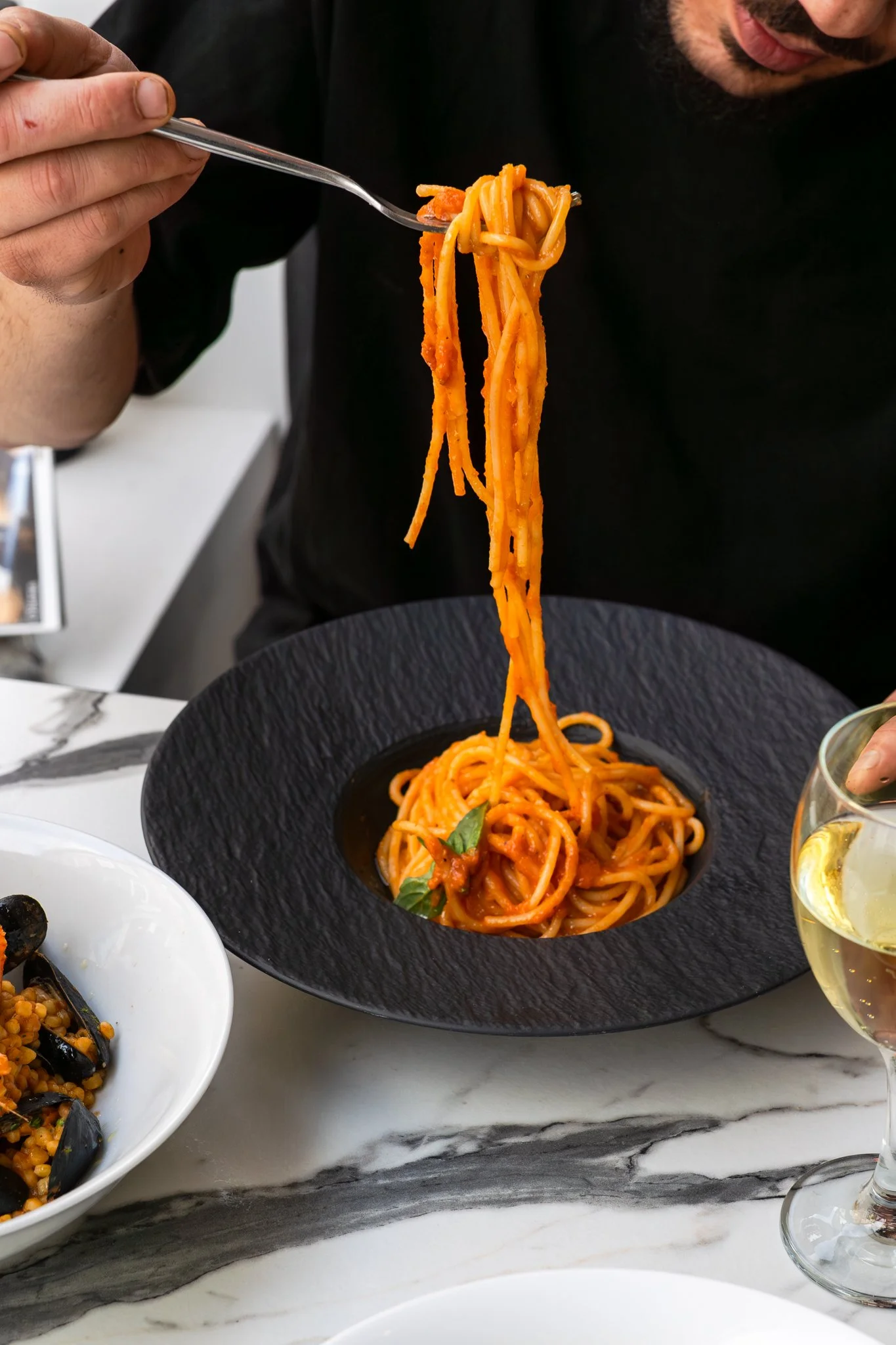 Person lifting spaghetti with tomato sauce on a fork above a black plate of pasta with basil leaves, on a white marble table with a glass of white wine nearby.