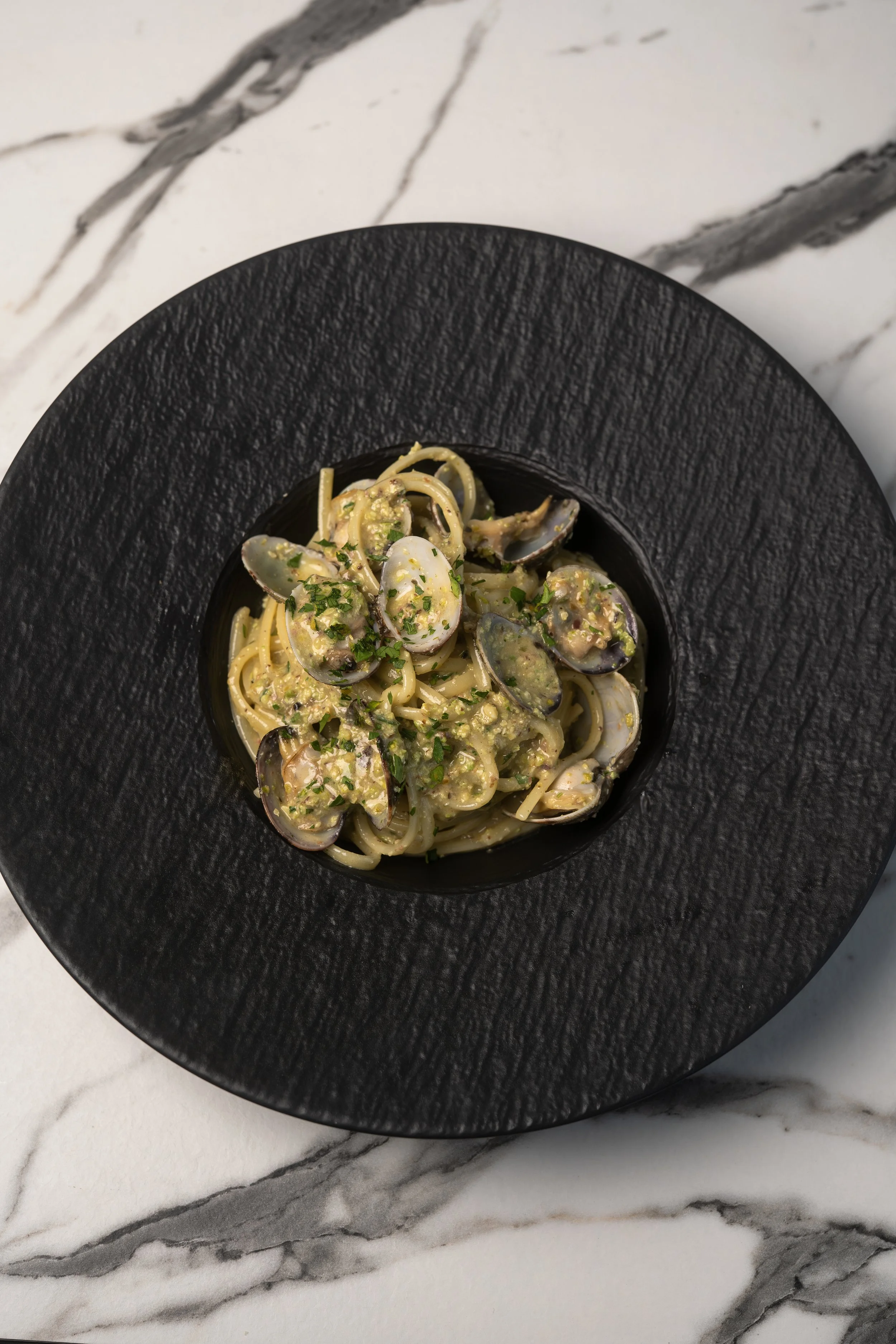 Spaghetti pasta with clams and herbs in a black bowl on a marble surface.