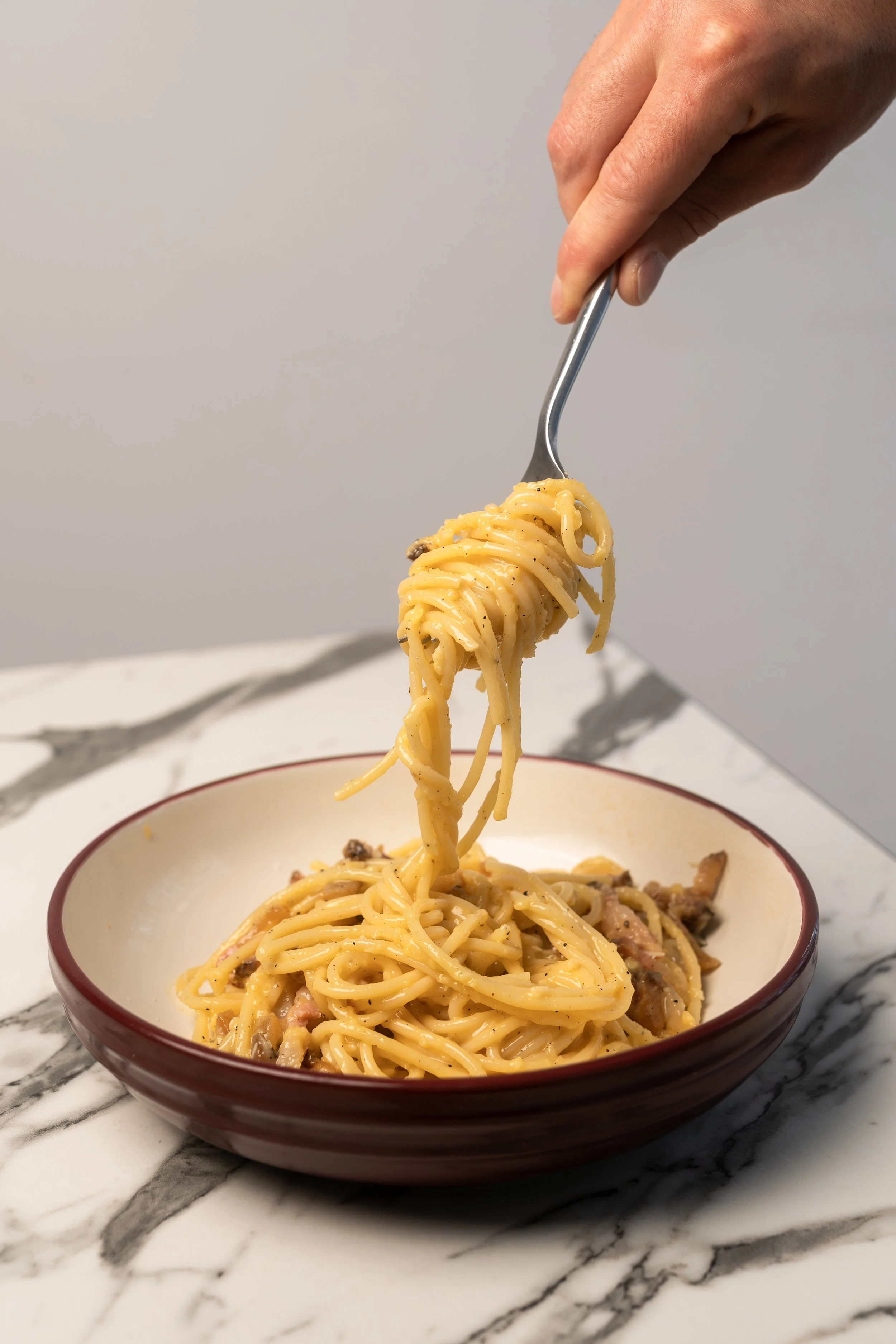A person is lifting a forkful of spaghetti with sauce and bits of meat from a bowl of spaghetti on a marble surface.