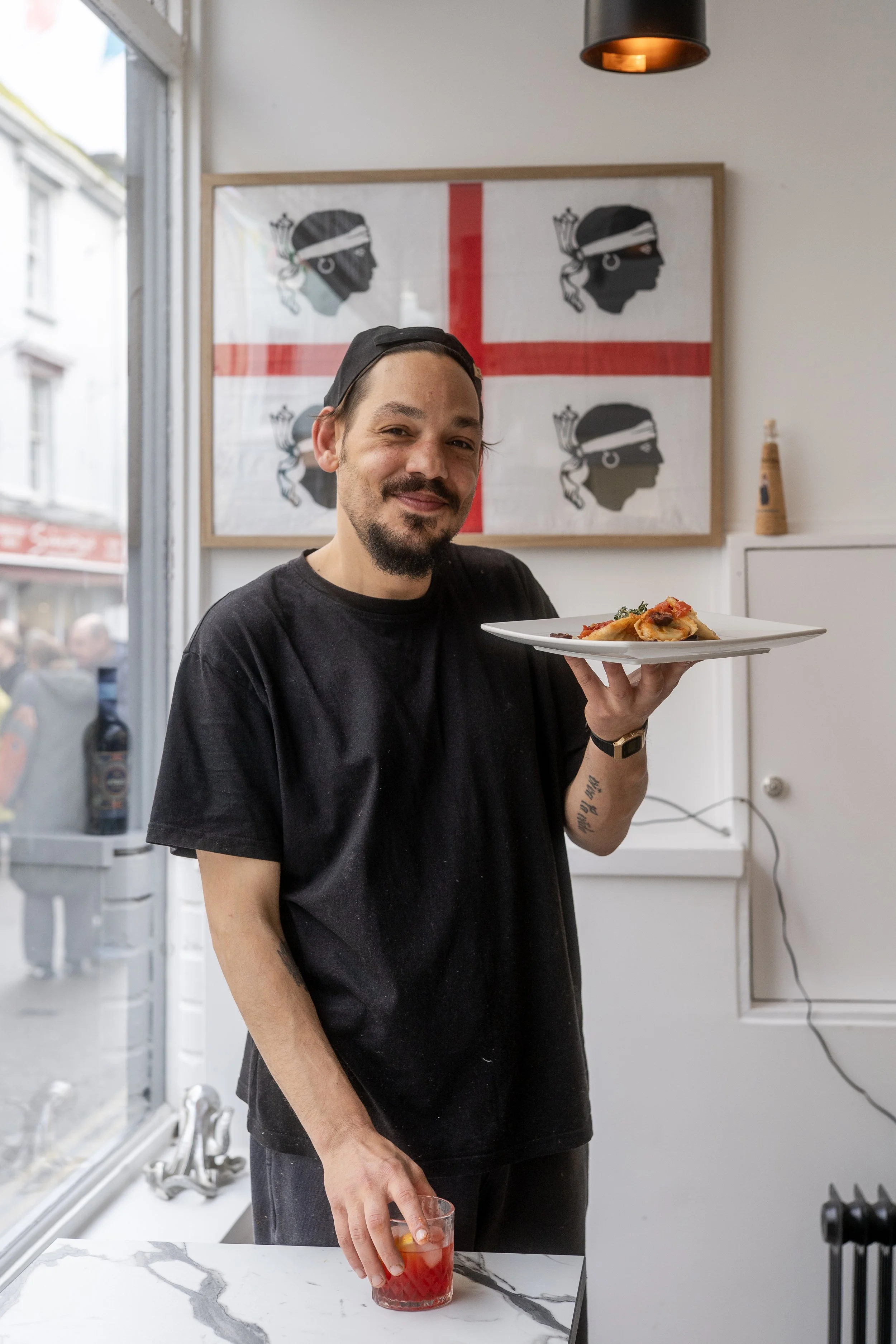 A man with a black t-shirt and a backwards baseball cap holding a white plate with food, standing inside a restaurant near a large window with a street view outside. There is artwork on the wall behind him and a drink with red liquid in his other hand.