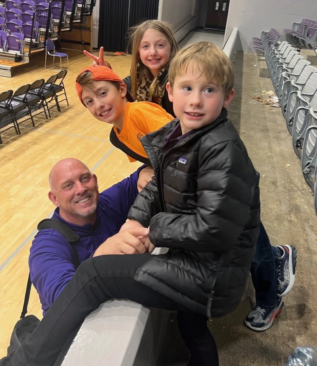 Four children and an adult in an indoor space, with chairs along the walls, smiling and engaging with each other.