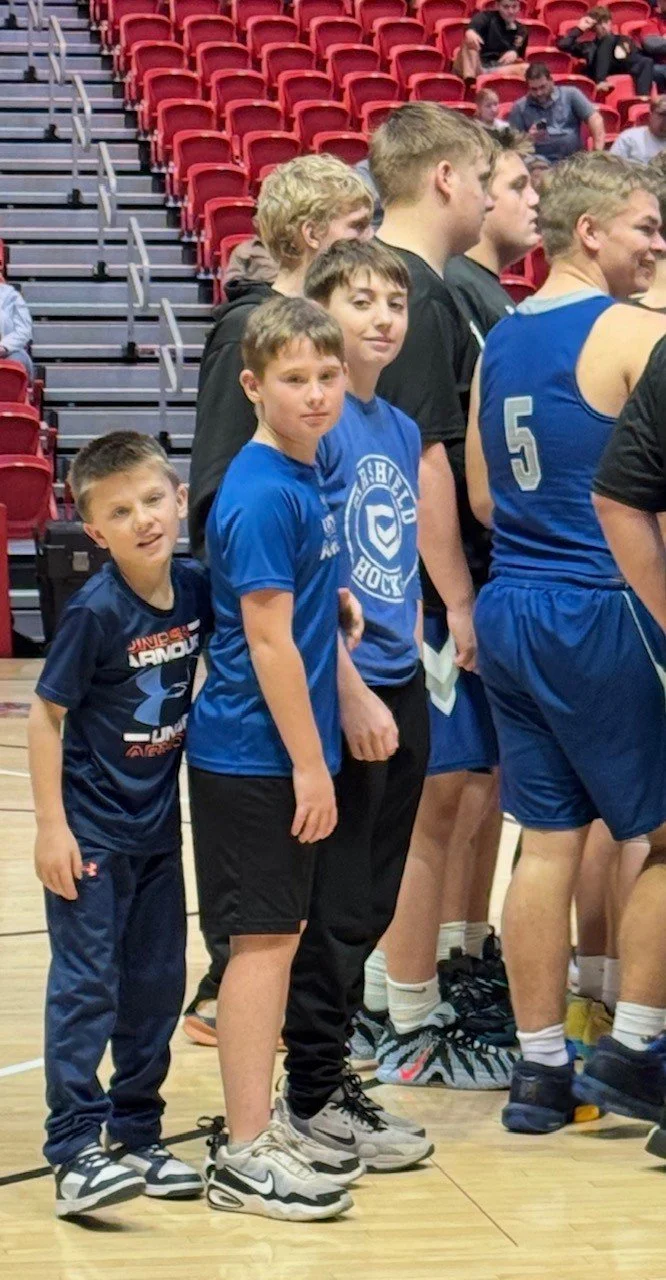 A group of young basketball players standing in a gymnasium, some wearing blue jerseys with a circular logo, others in black and blue shorts, with red bleachers in the background.