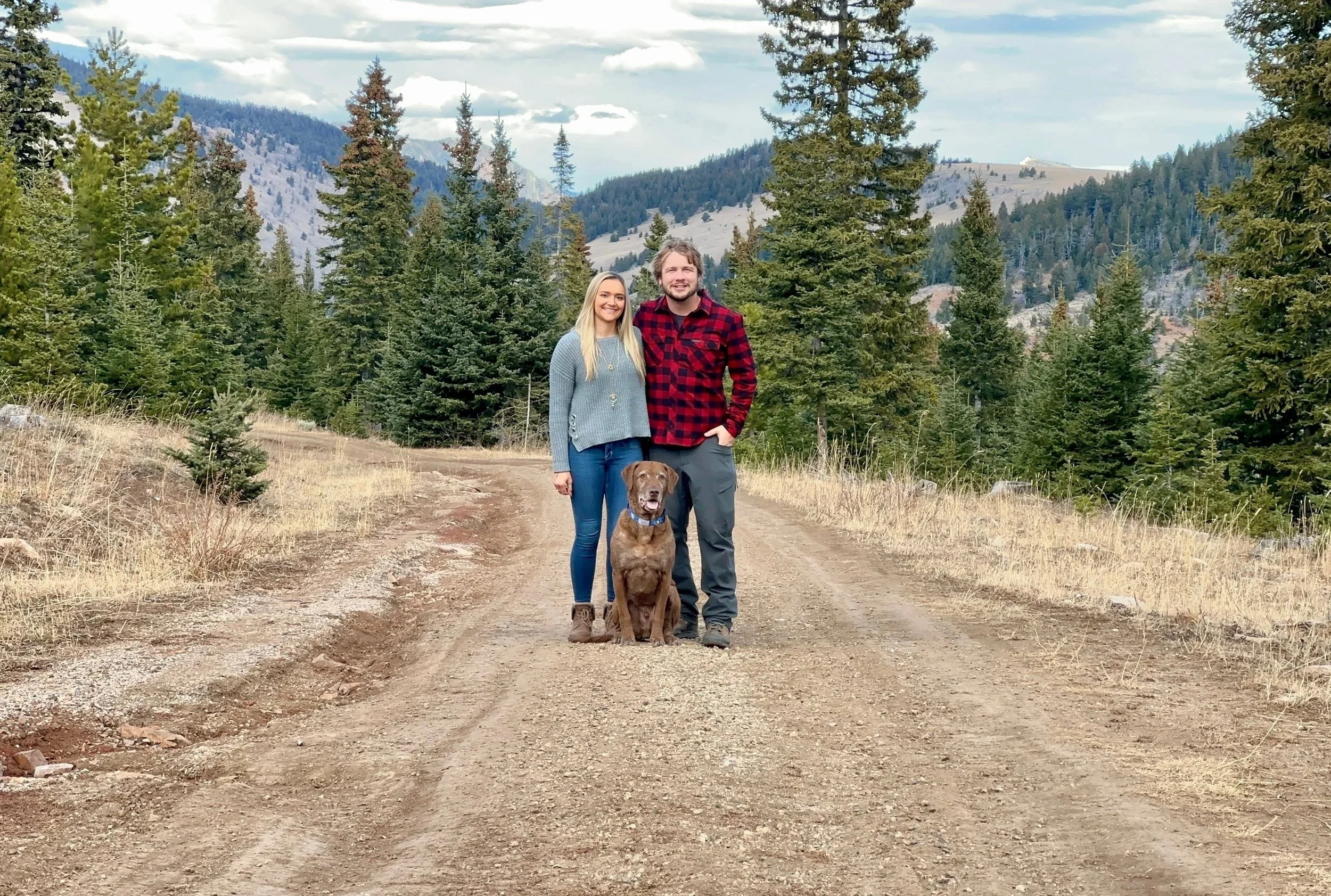 A couple with a brown dog standing on a dirt trail in a forested mountainous area.
