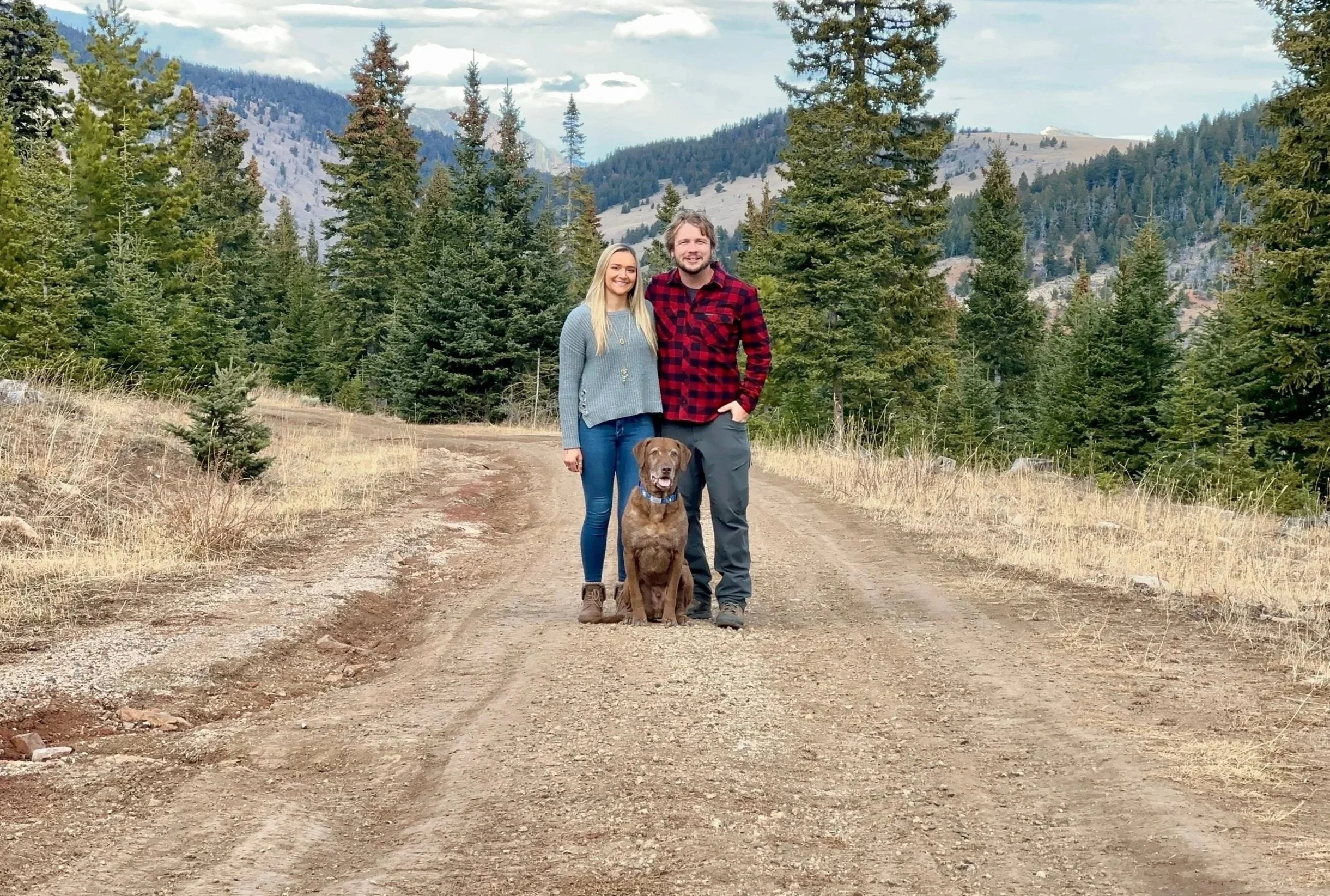 A couple and their dog standing on a dirt trail in a mountainous forested area with evergreen trees and cloudy sky.