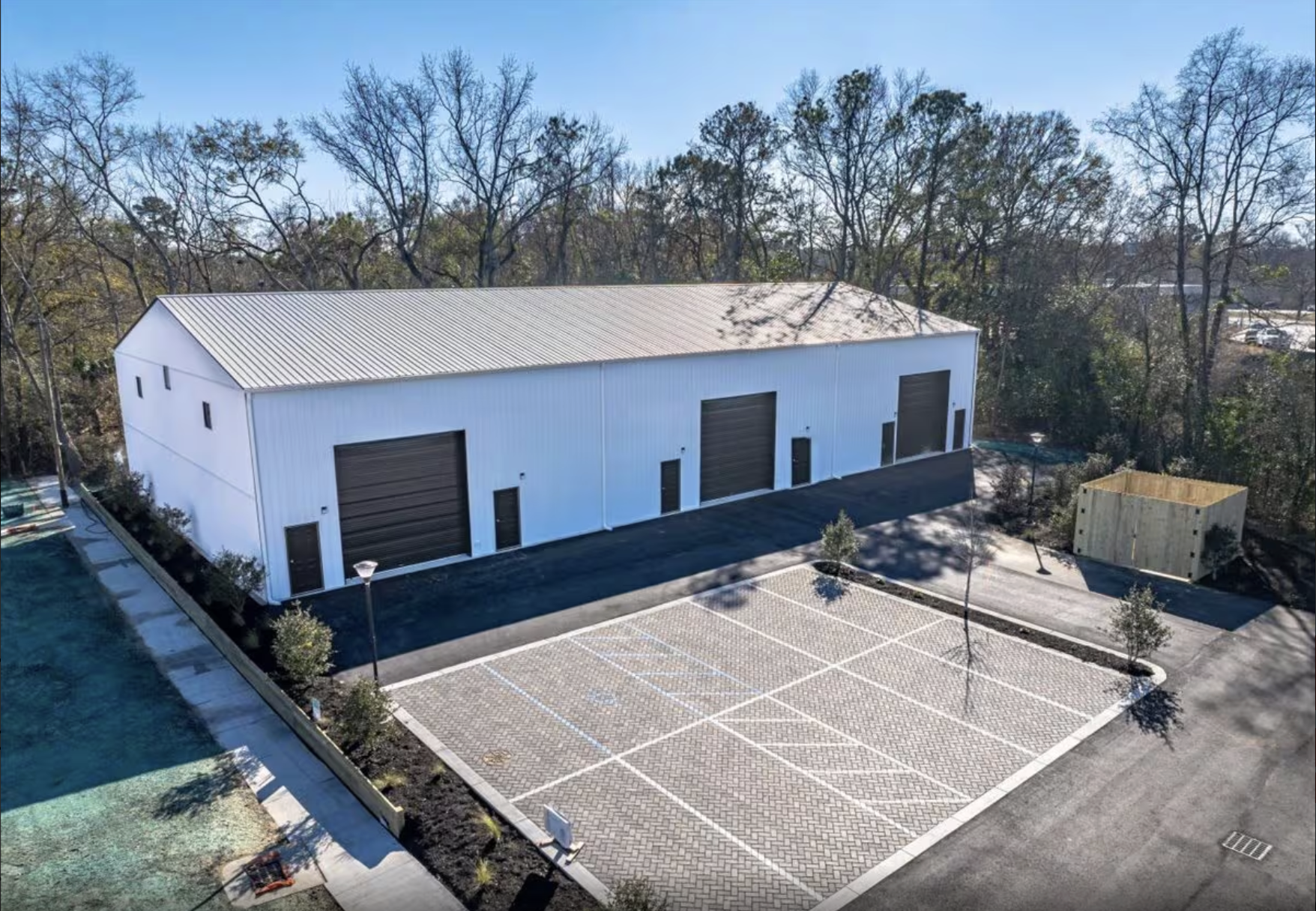 A large white industrial building with three roll-up garage doors, surrounded by a parking lot with empty spaces, small trees, and a wooded area in the background.