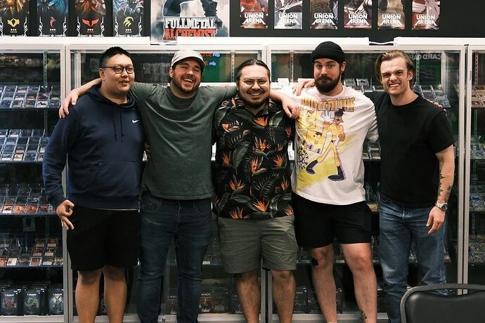 Five young men standing together inside a store with shelves of DVDs or video games behind them, smiling and posing for the photo.