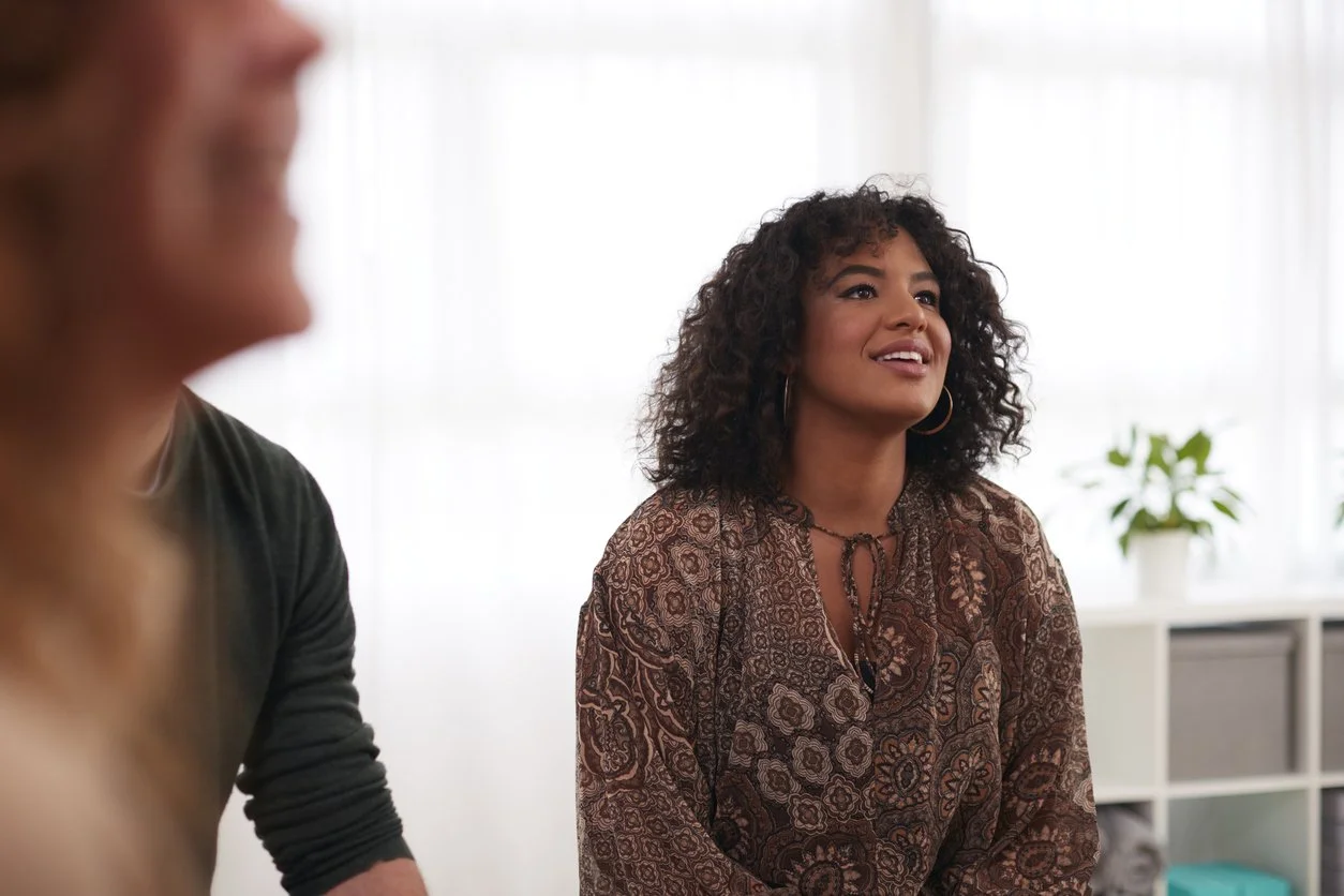 A woman with curly hair wearing a patterned blouse sitting and smiling in a bright room with a plant in the background.