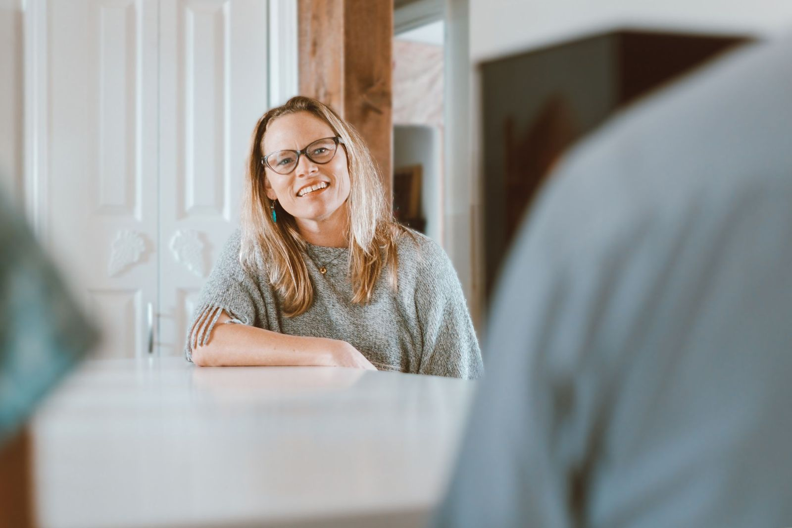 A woman with glasses and long hair, smiling, sitting at a table in a cozy indoor setting.