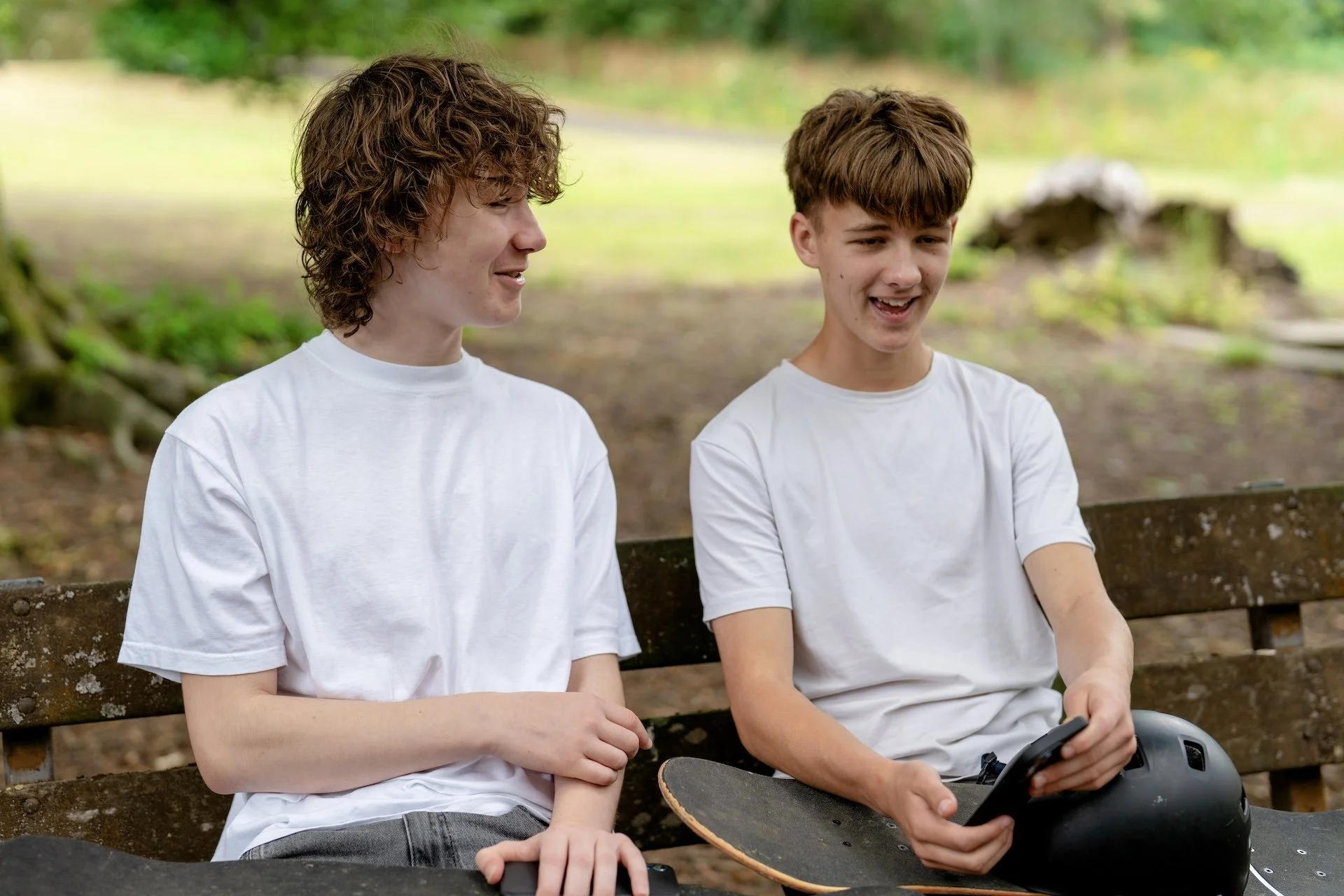 Two teenage boys with curly and straight brown hair sitting on a park bench, smiling and talking, one holding a skateboard, the other holding a smartphone, in a green outdoor setting.