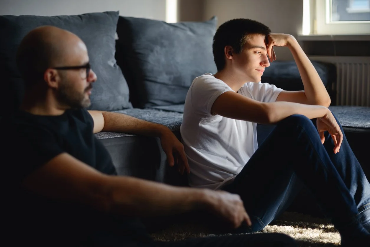 Two men sitting on the floor in front of a sofa, one in profile with glasses and a beard, and the other looking out a window with sunlight on his face, in a room with a gray sofa and a window.