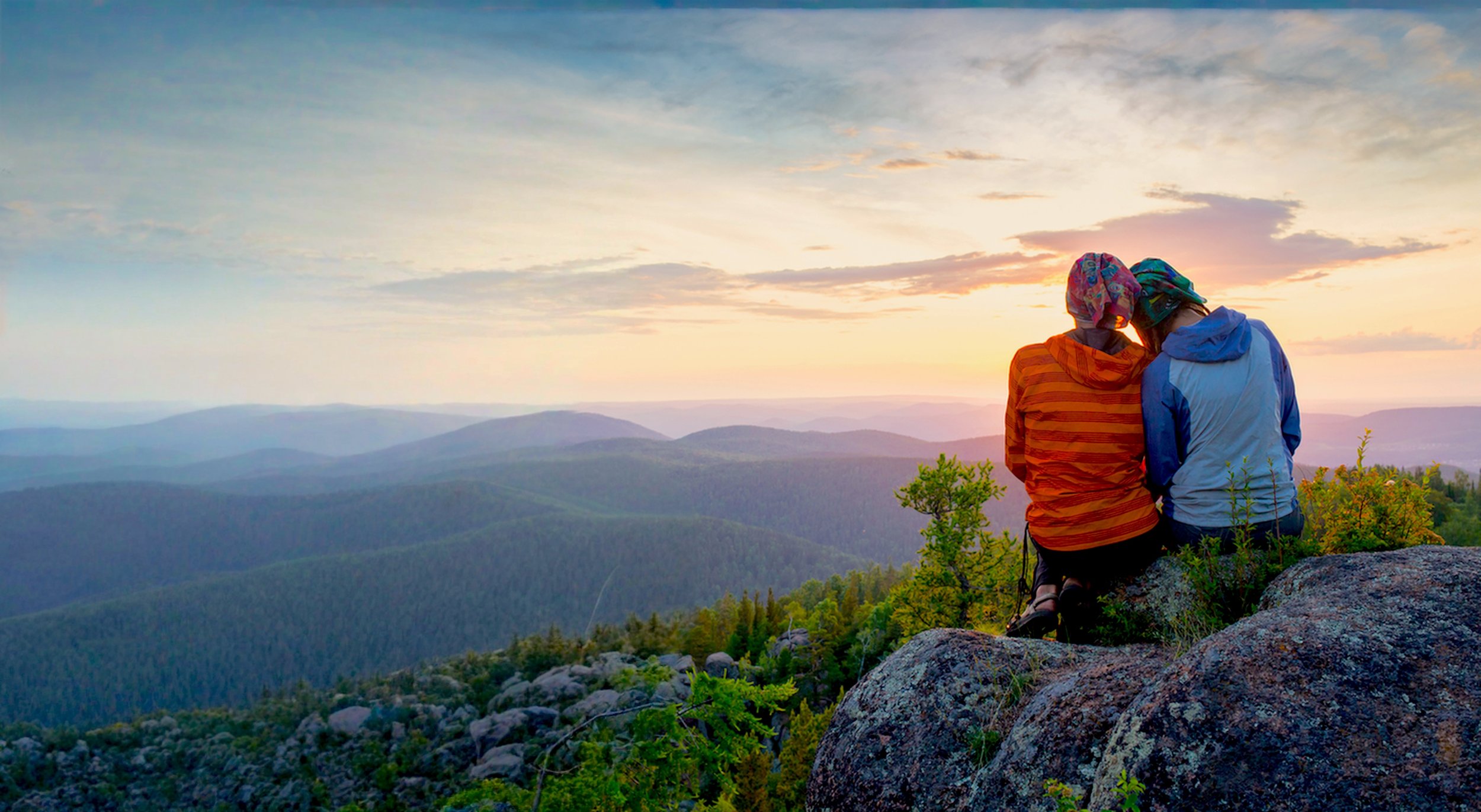 Two people sitting on rocks, watching a sunset over a mountainous landscape with trees and a cloudy sky.