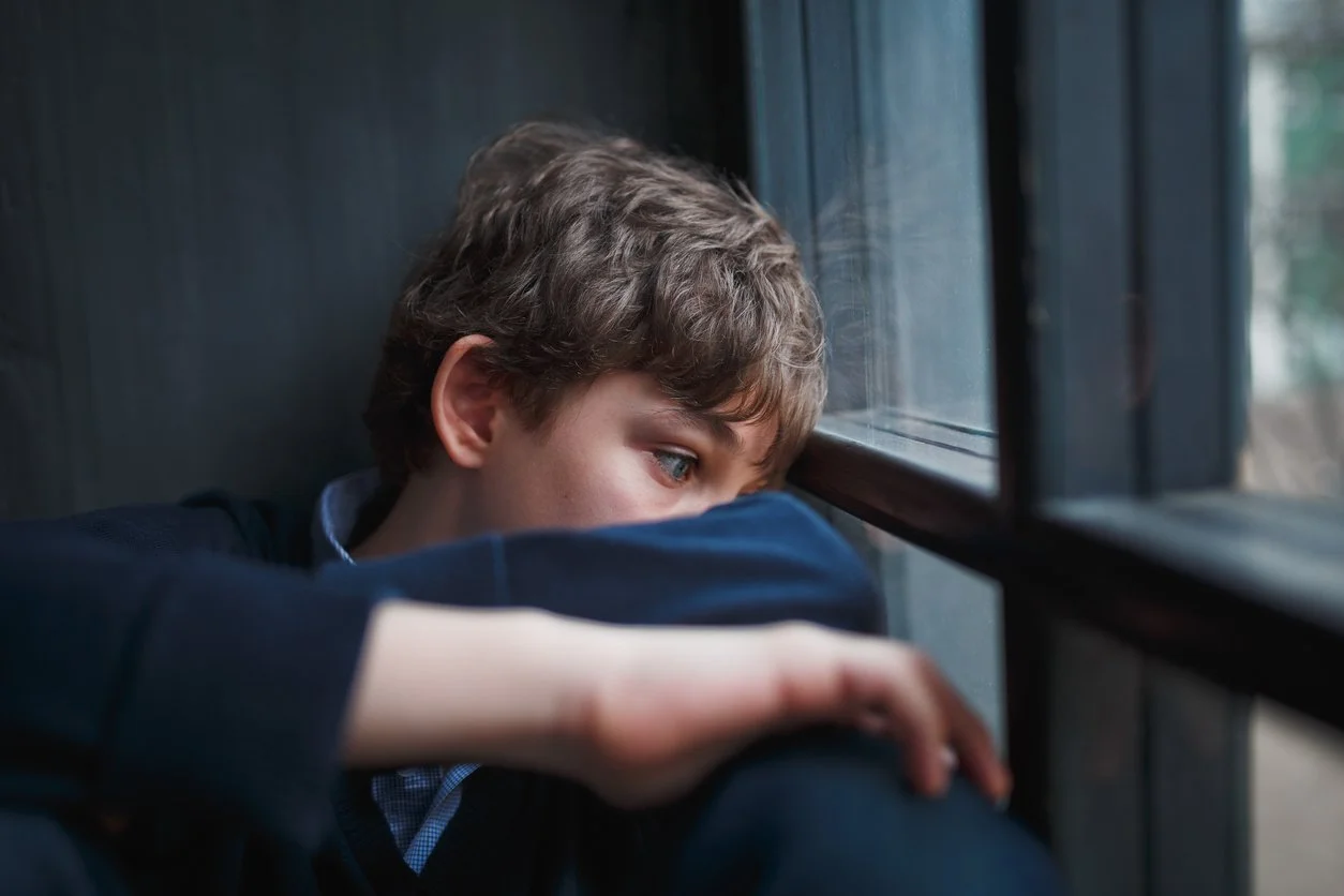 Young boy looking out a window with a sad expression, resting his arms on his knees in a dimly lit room.