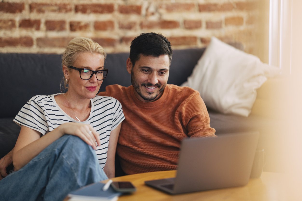 A man and woman sitting on a couch, looking at a laptop together, smiling. The woman has blonde hair, glasses, and is wearing a striped shirt. The man has dark hair, a beard, and is wearing an orange sweater.