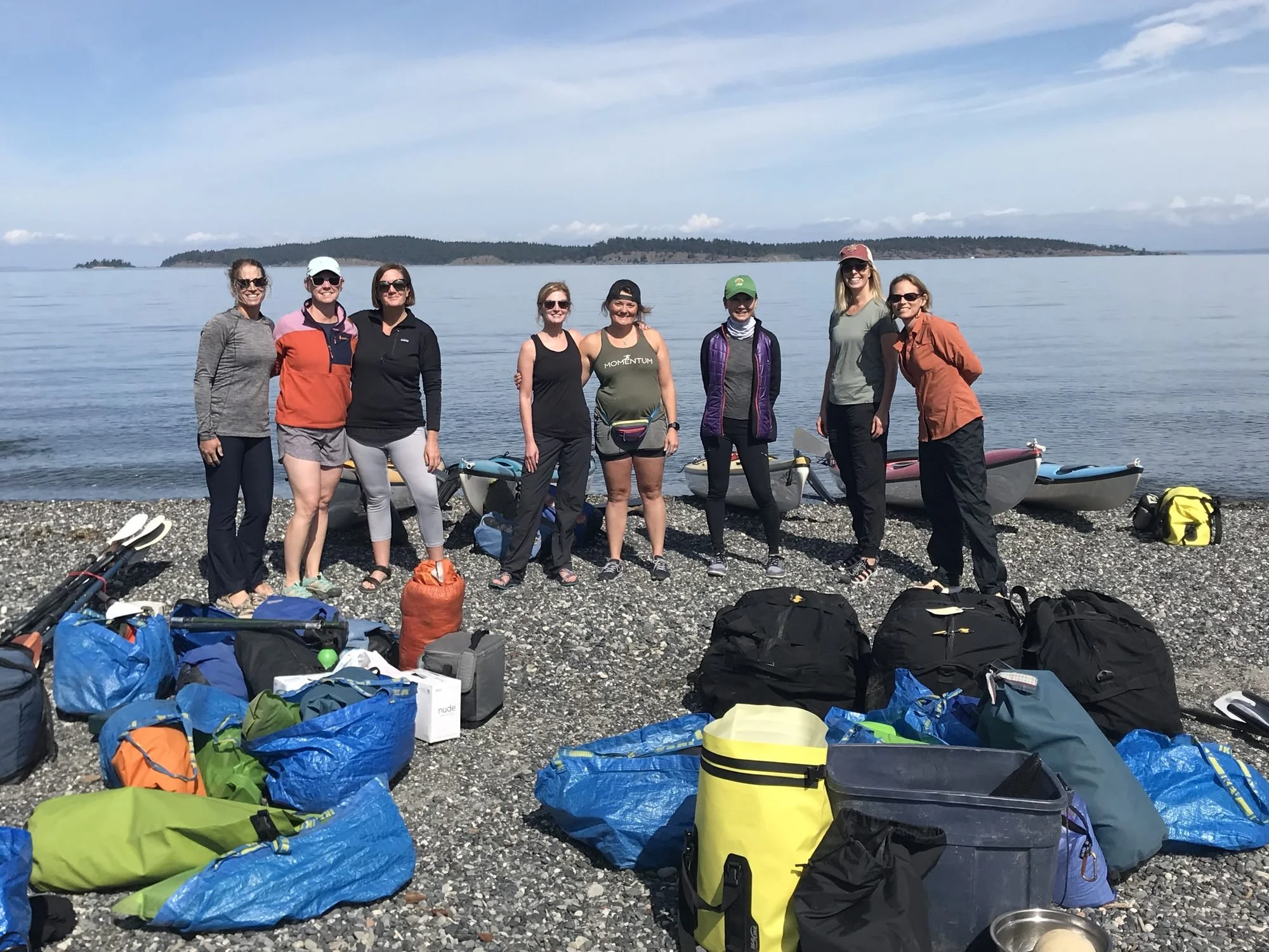 A group of eight women standing on a rocky shoreline after a kayaking trip, with kayaks and gear arranged in front of them, overlooking a calm body of water and distant islands under a partly cloudy sky.
