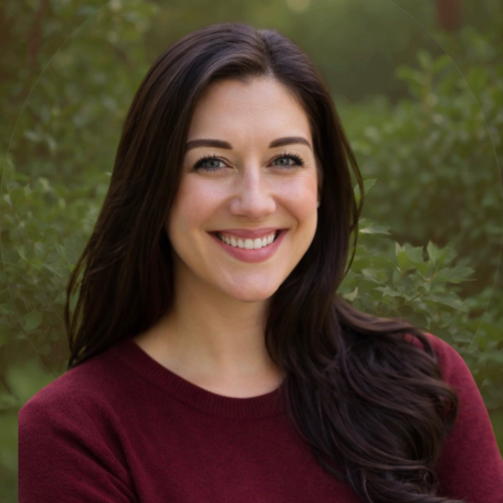 A woman with long dark hair smiling outdoors with green foliage in the background.