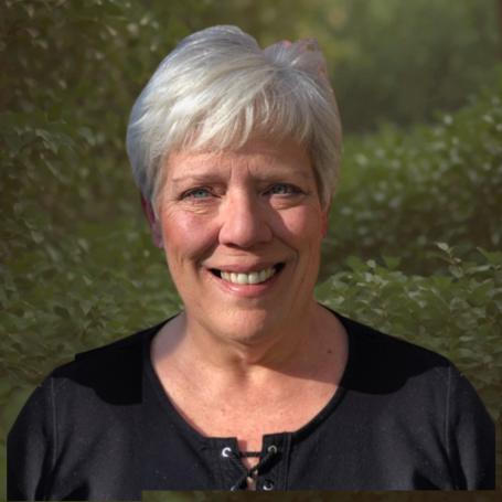 A smiling elderly woman with short white hair standing outdoors with green foliage in the background.